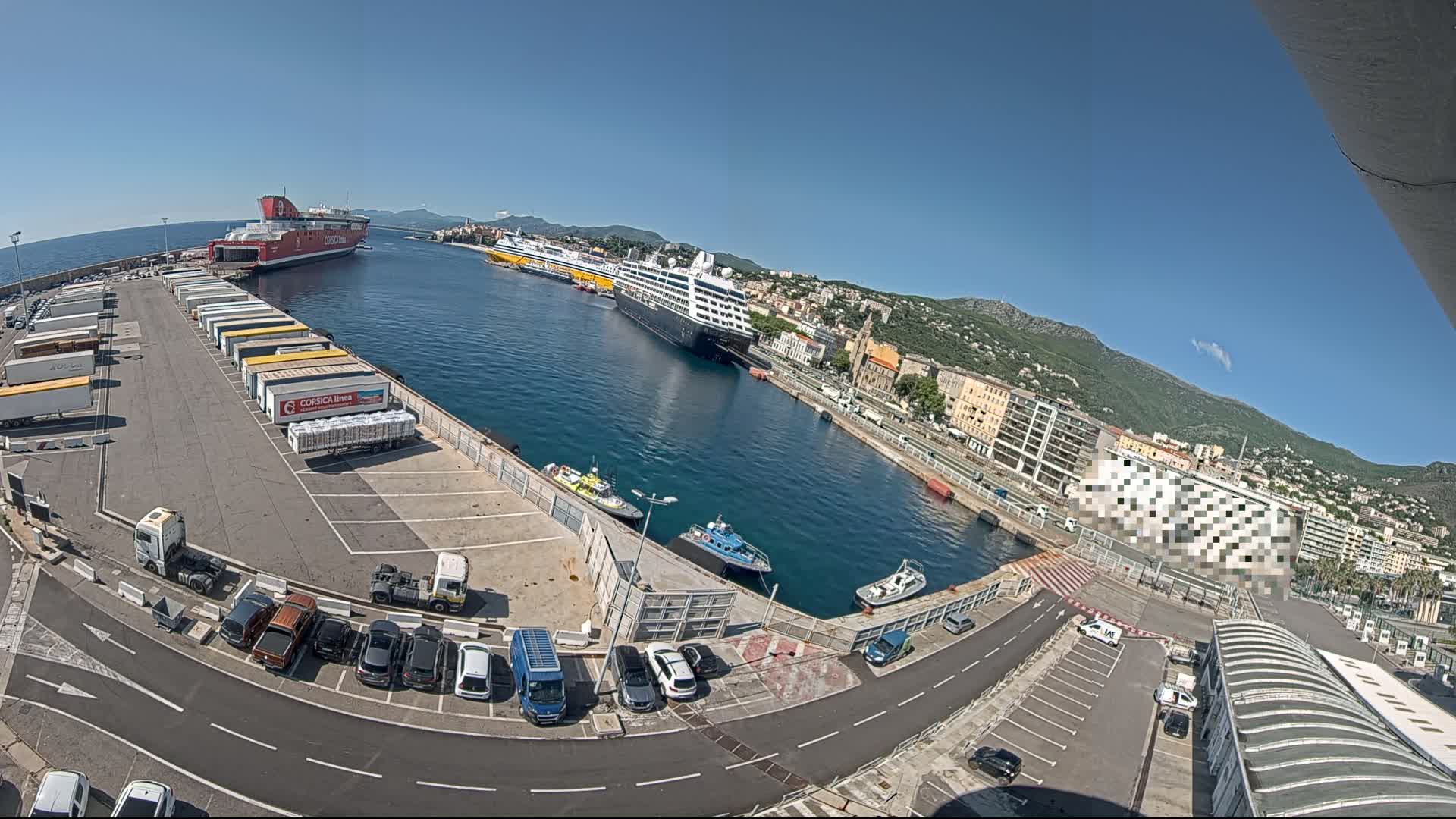 A sunny harbor scene shows several docked ferries and cruise ships, numerous parked cars and trucks, and several small boats.