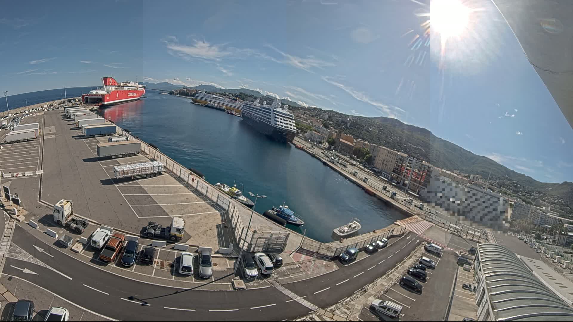 A sunny day at a harbor shows several large ships docked, with cars and trucks parked in a lot nearby.