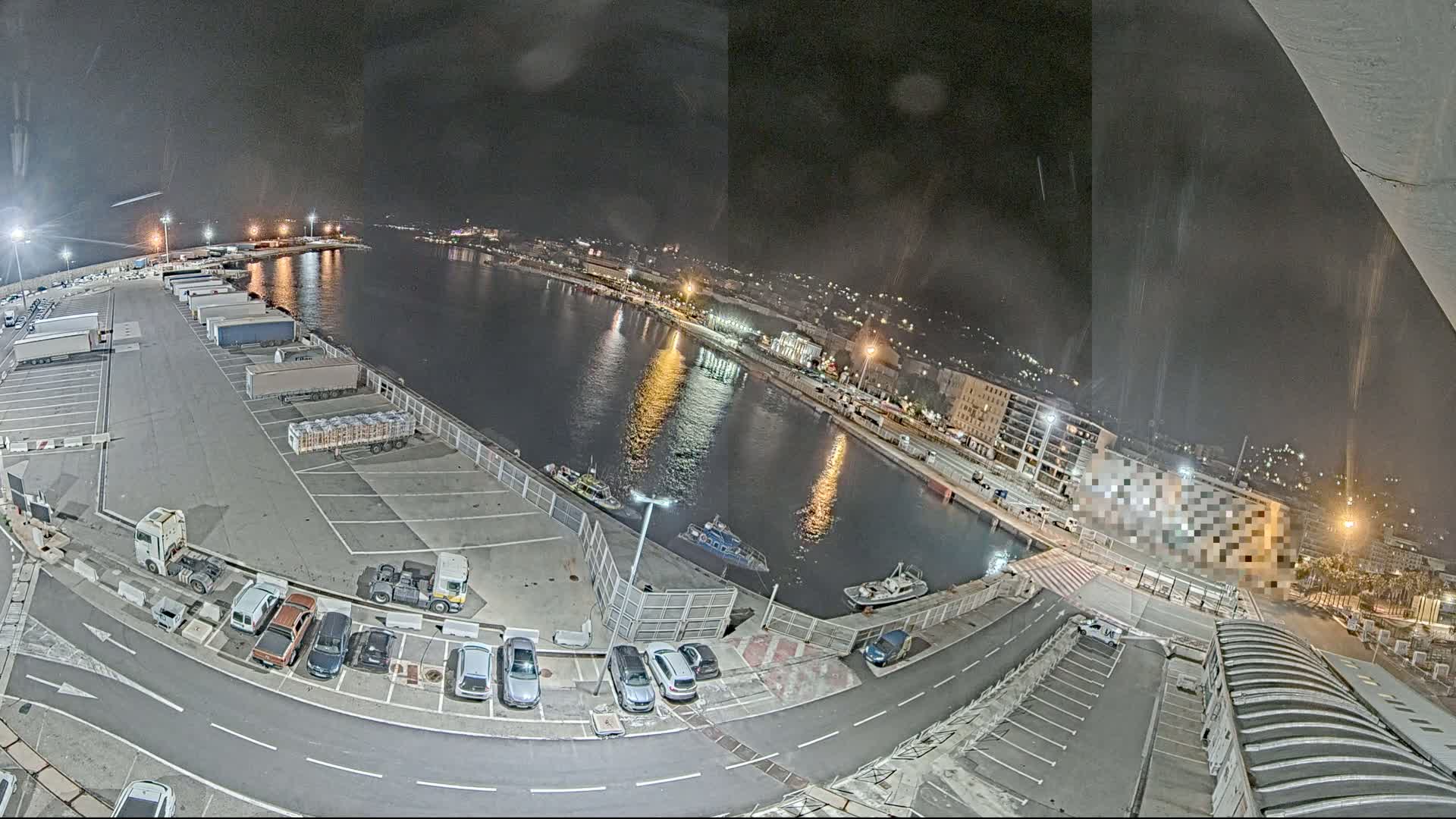 A nighttime, high-angle view of a harbor with numerous trucks and cars parked in lots, several small boats on the water, and city lights visible in the distance under a slightly hazy sky.
