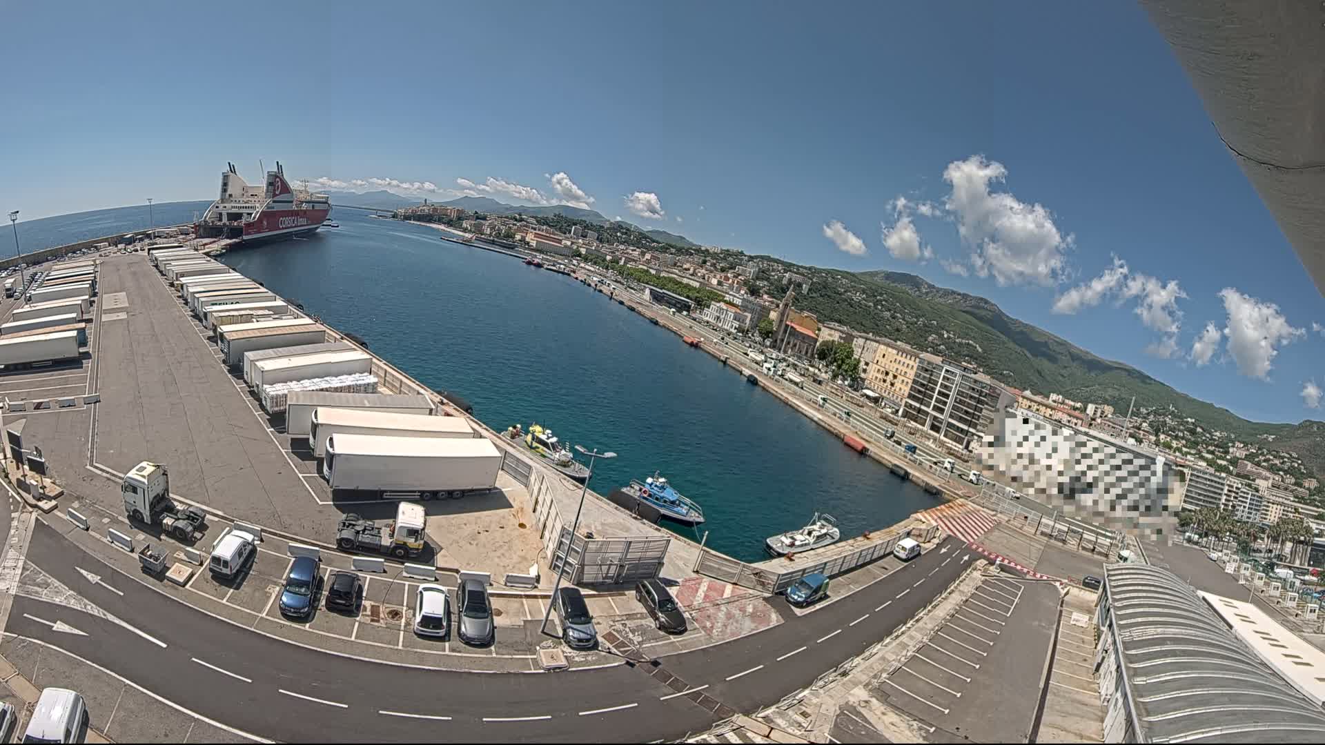A large ferry is docked at a port on a sunny day with a few scattered clouds;  the port area shows numerous parked trucks and cars, and small boats are in the water.