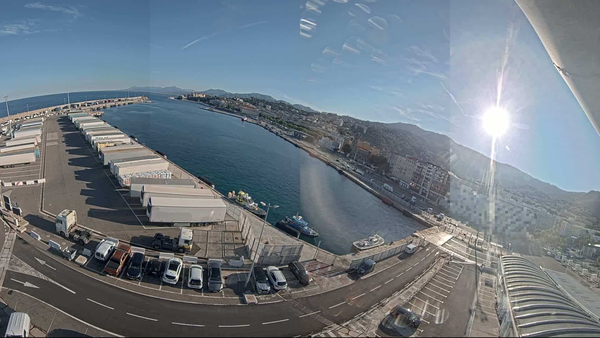 A sunny day at a harbor shows a large parking lot filled with semi-trucks and cars, next to a waterway with several small boats and a city in the background.