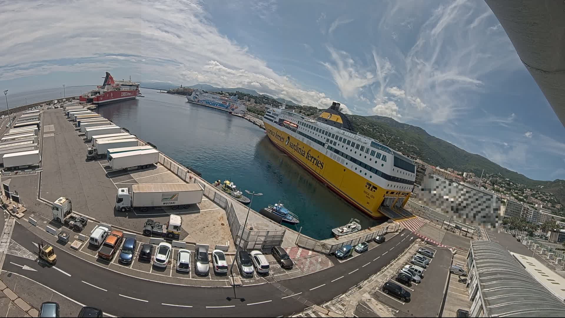 A sunny day at a harbor shows several ferries docked, numerous trailers parked in a lot, and cars in a parking area.