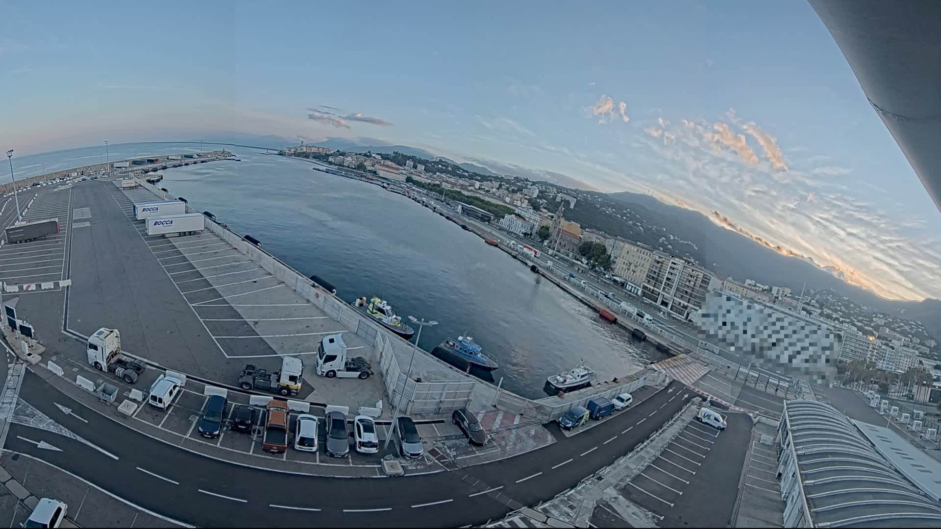 A high-angle, wide shot shows a harbor with numerous cars and trucks parked in lots, several small boats in the water, and a city in the background under a mostly clear, light blue sky with some high clouds.