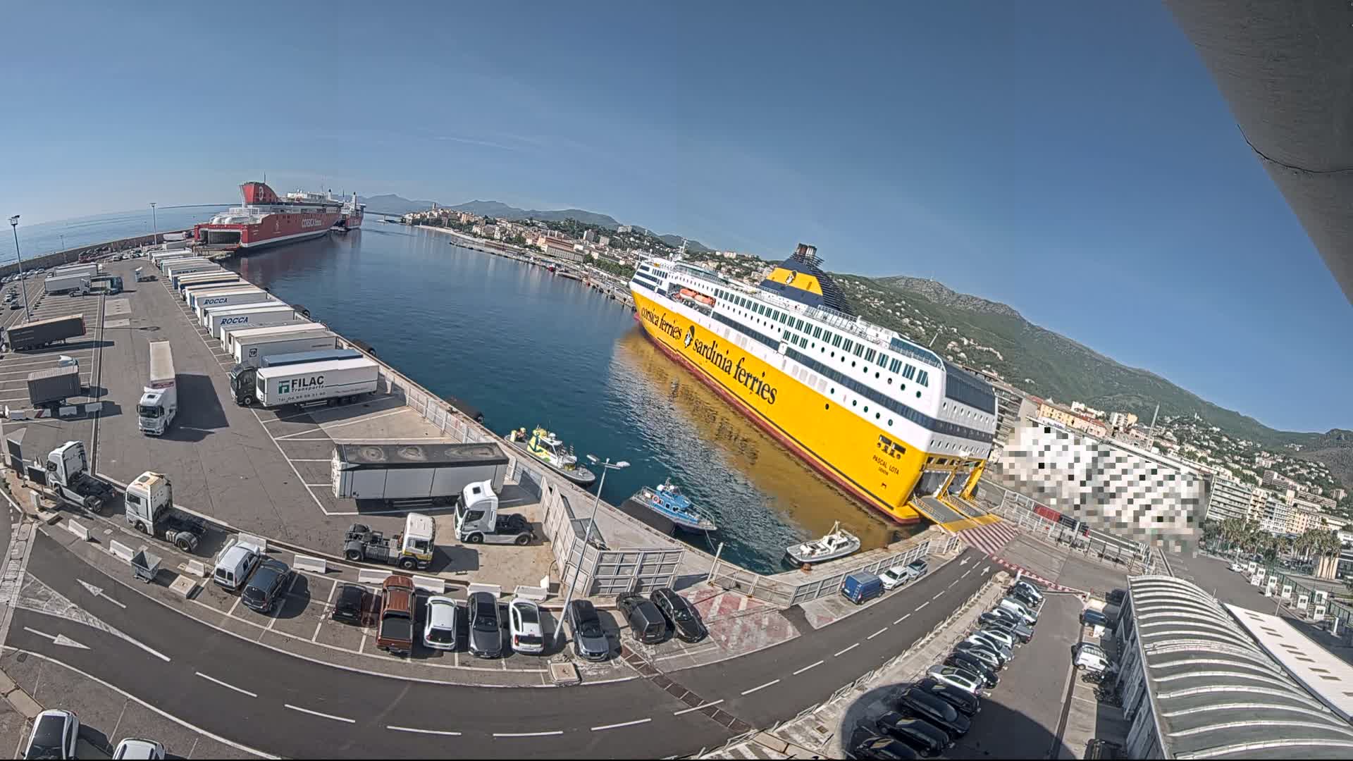 A large yellow and white ferry is docked at a port on a sunny day, surrounded by trucks, cars, and smaller vessels; mountains are visible in the background.