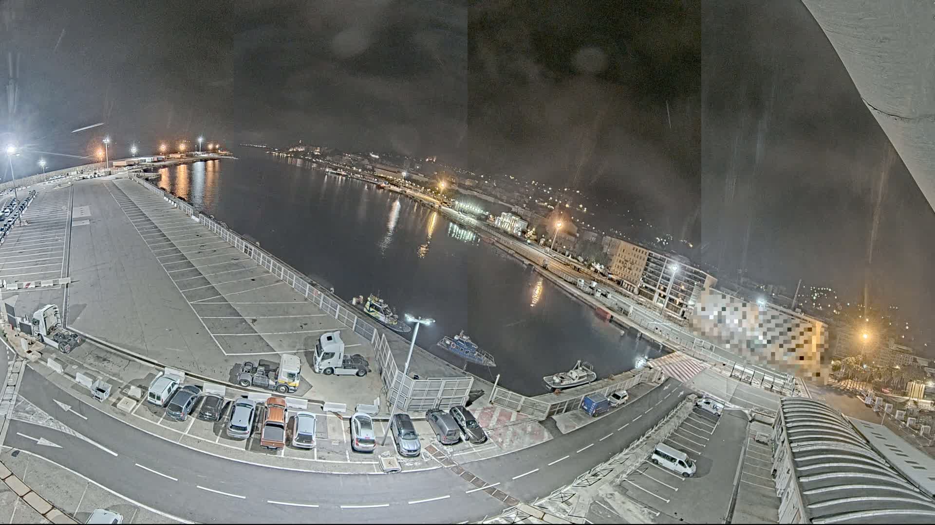 A nighttime, high-angle view of a harbor with a large empty parking lot, several vehicles, and a few boats on the water.
