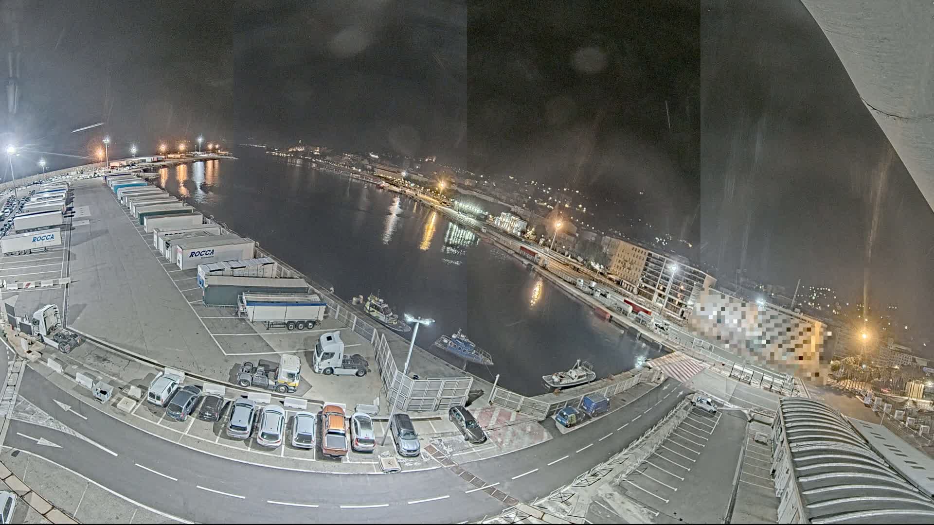A nighttime aerial view of a harbor with parked cars and trucks along a waterfront, with several small boats visible in the water.