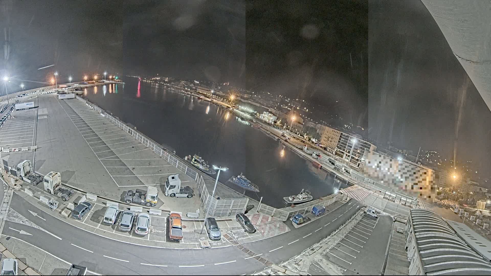 A nighttime aerial view of a harbor shows a mostly empty parking lot next to a calm body of water with several boats and buildings along the shore under a dark, slightly hazy sky.