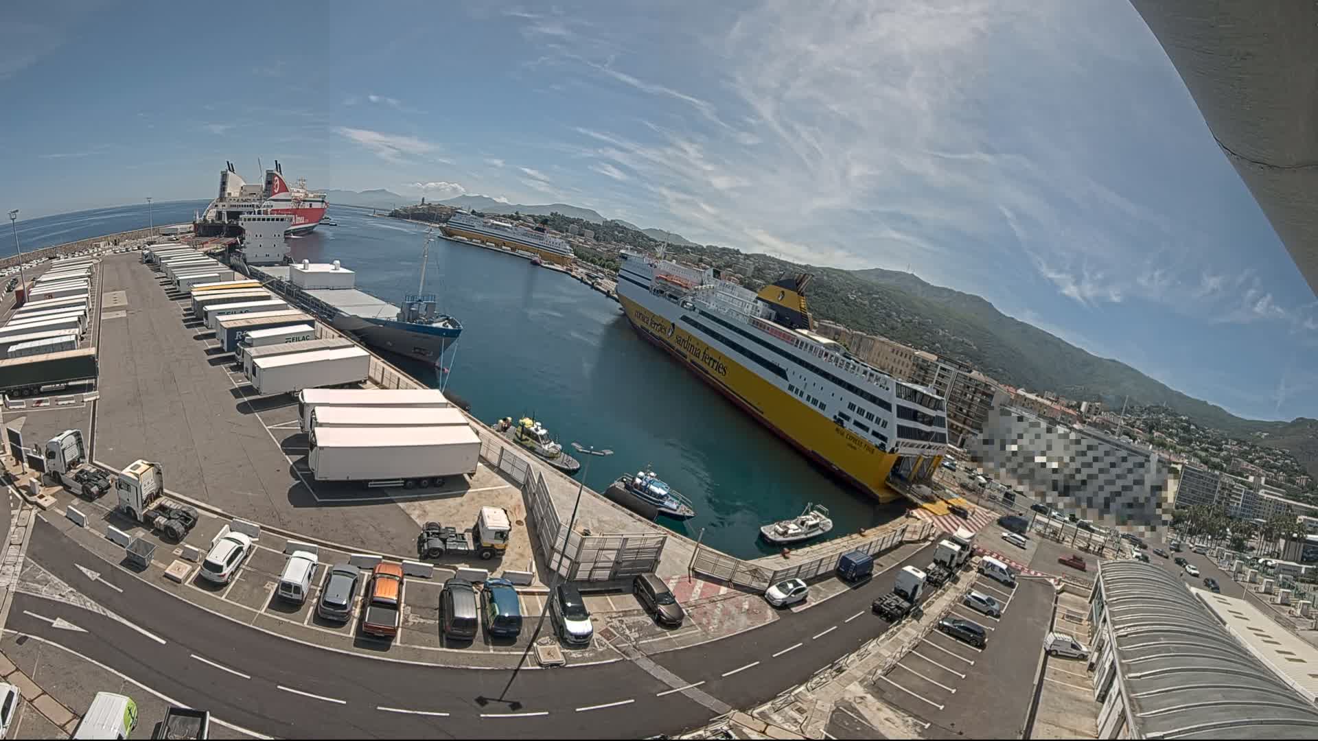 A ferry terminal on a sunny day shows several ferries docked, numerous trailers parked, and cars in a nearby lot, with mountains visible in the distance.
