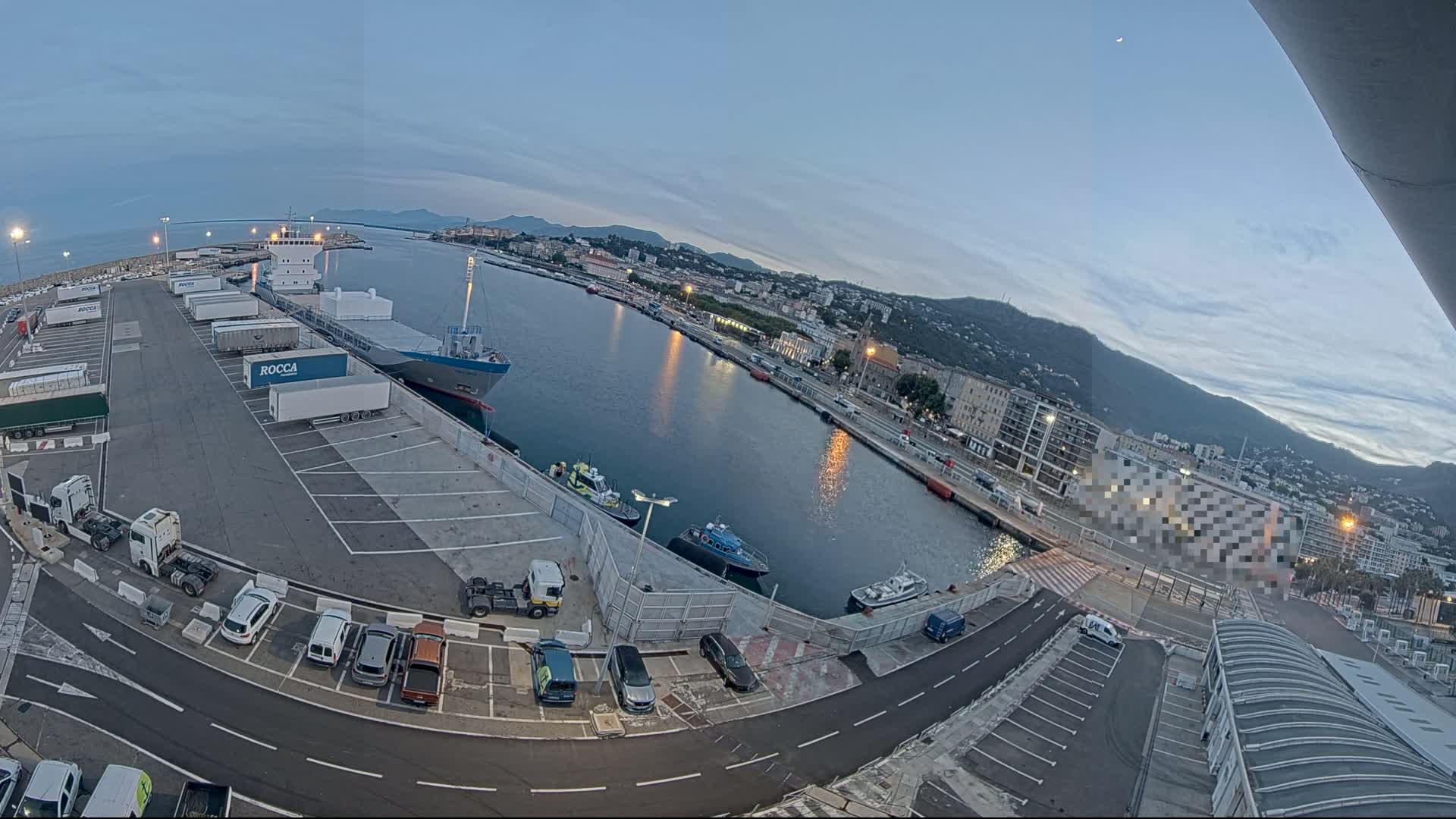 A cargo ship is docked at a harbor next to a parking lot filled with cars and trucks under a twilight sky.