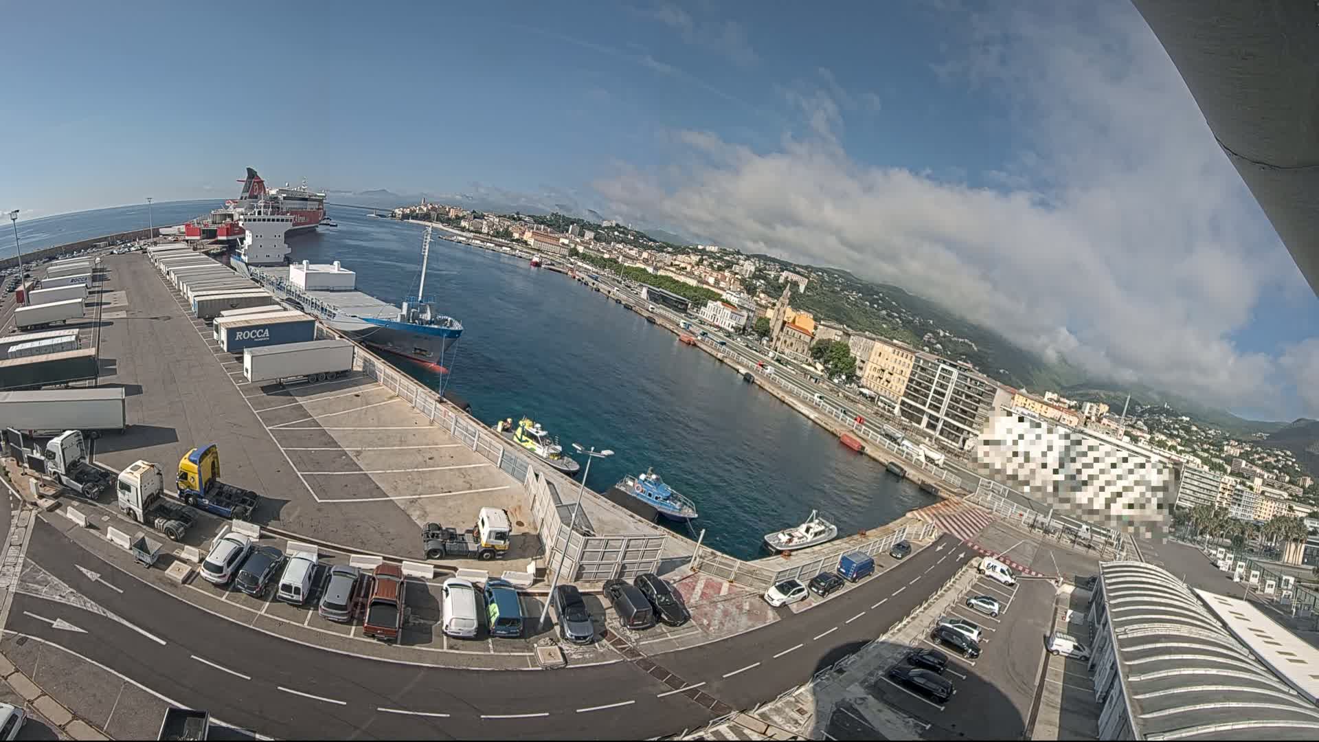 A partly cloudy day overlooking a harbor with docked ships, cargo containers, and parked cars, and a city in the background.