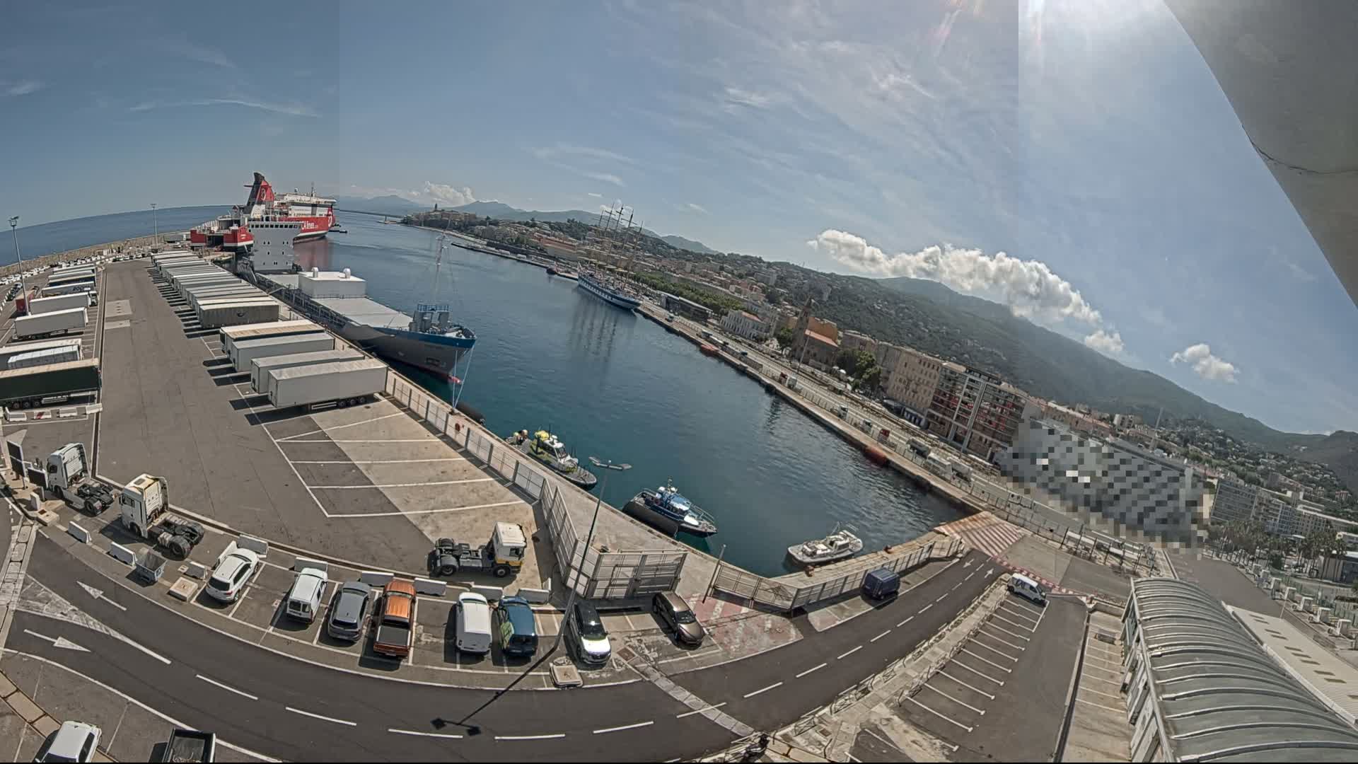 A sunny day at a harbor shows cargo ships, smaller boats, and a parking lot filled with cars and trucks, all situated beside a waterfront city with mountains in the background.
