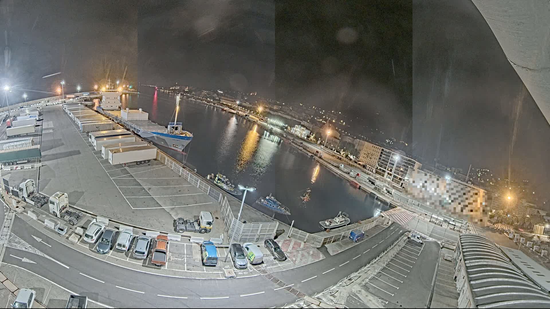 A nighttime aerial view of a harbor with a cargo ship docked, numerous trailers and cars parked, and several small boats on the water.