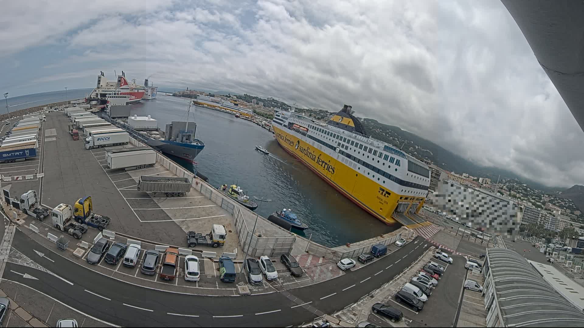A large yellow ferry is docked at a port under a cloudy sky, with several other ships, trucks, and cars visible in the area.