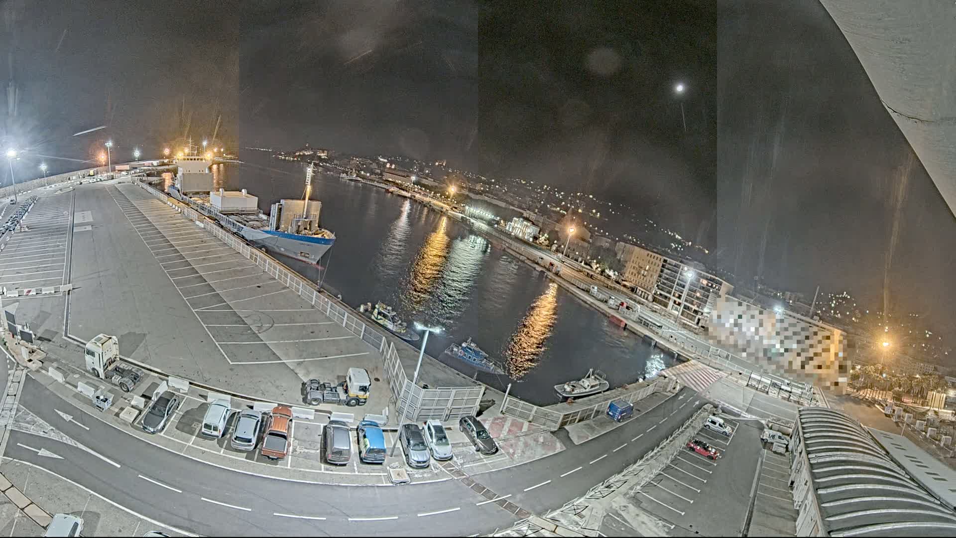 A nighttime aerial view of a harbor with a cargo ship docked, several smaller vessels on the water, and cars parked in a lot next to the water's edge under a clear, moonlit sky.