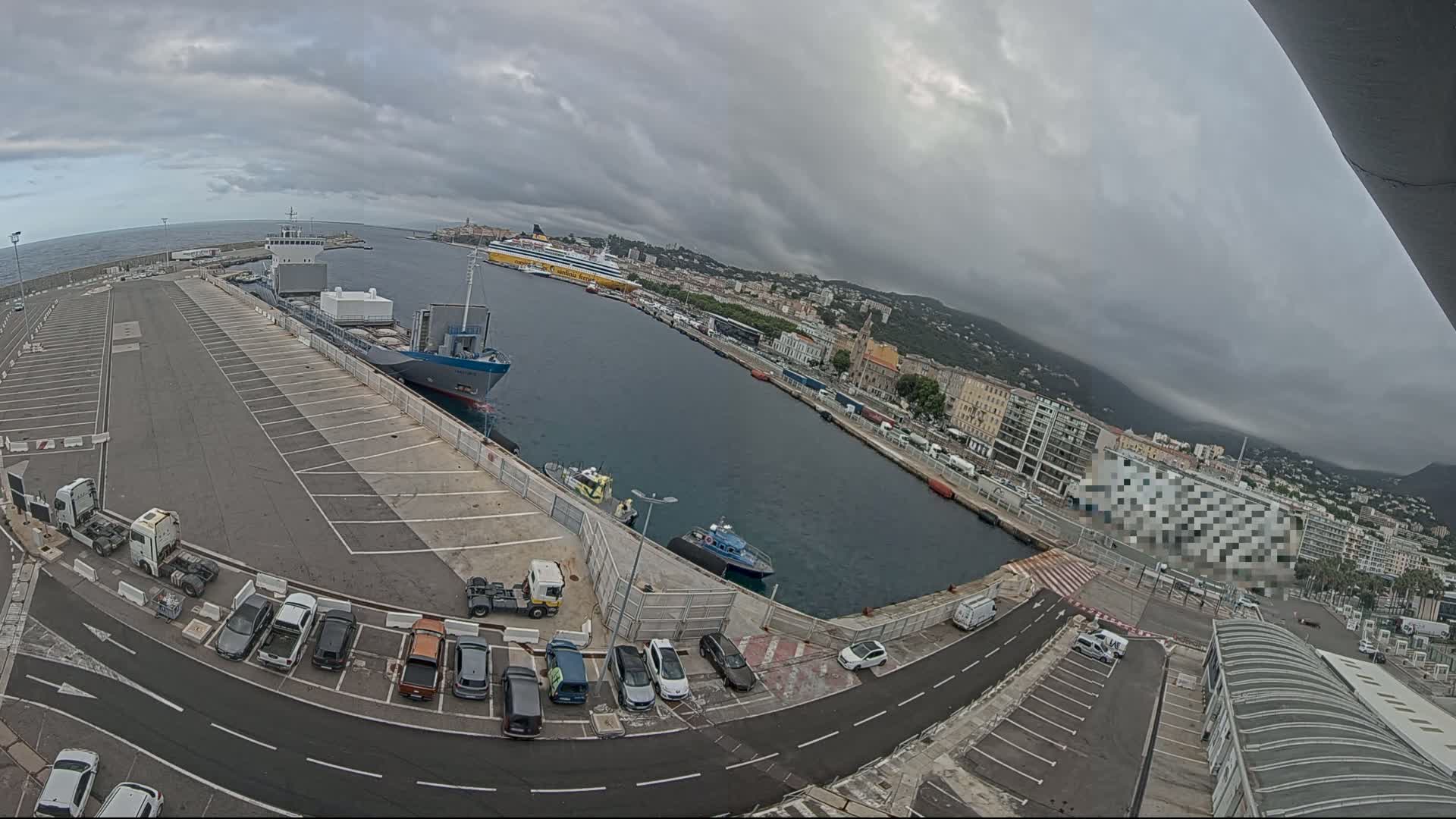 A harbor with overcast skies shows several docked ships, a parking lot filled with cars and trucks, and a city in the background.