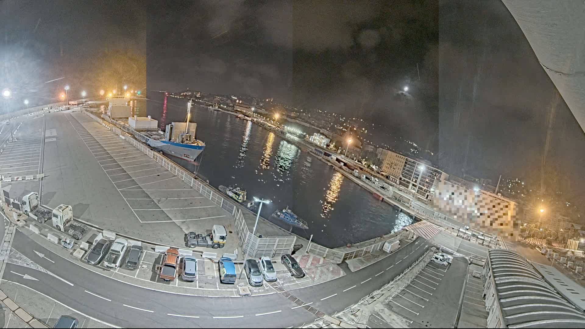 A nighttime, high-angle view of a harbor with a cargo ship docked, several small boats on the water, cars parked in lots, and city lights in the background.