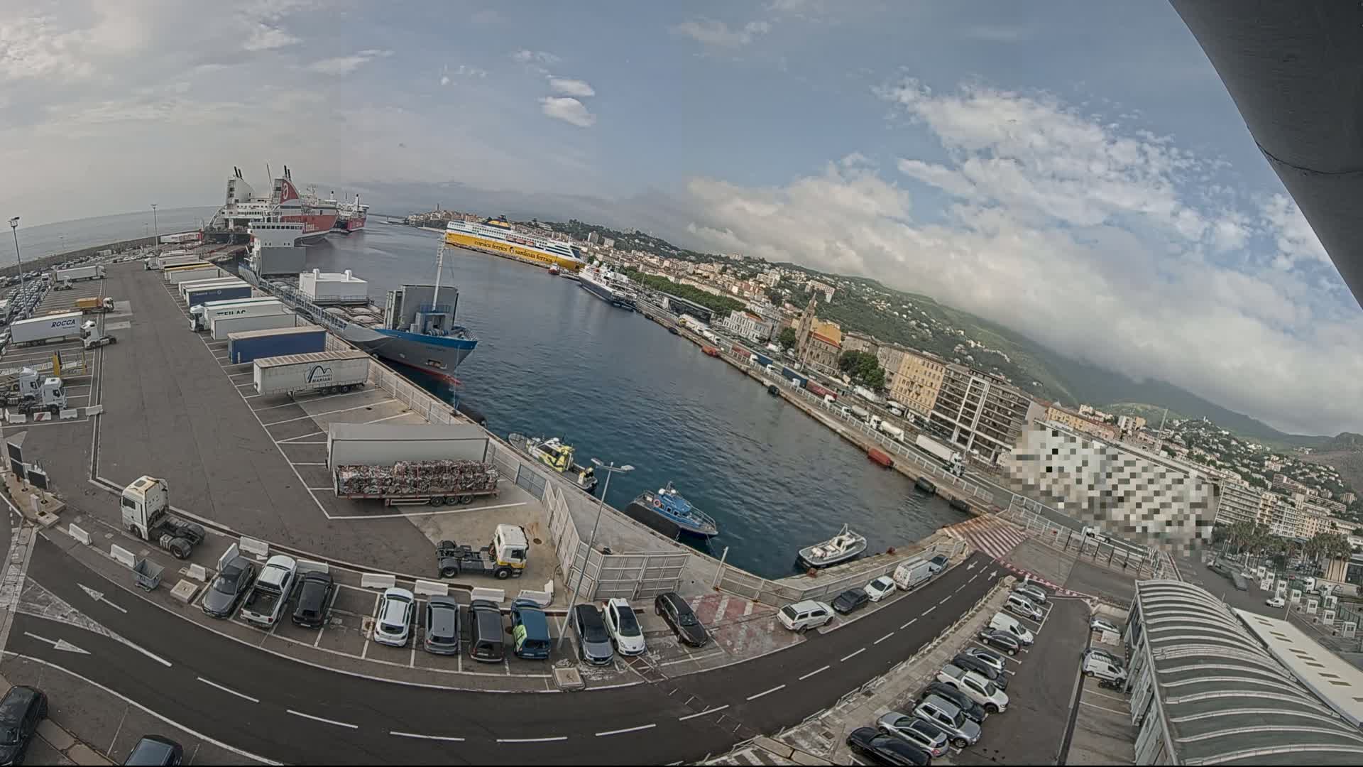 A partly cloudy day at a busy port shows several large ferries and cargo ships docked, numerous trucks and cars parked in lots, and smaller vessels in the water.