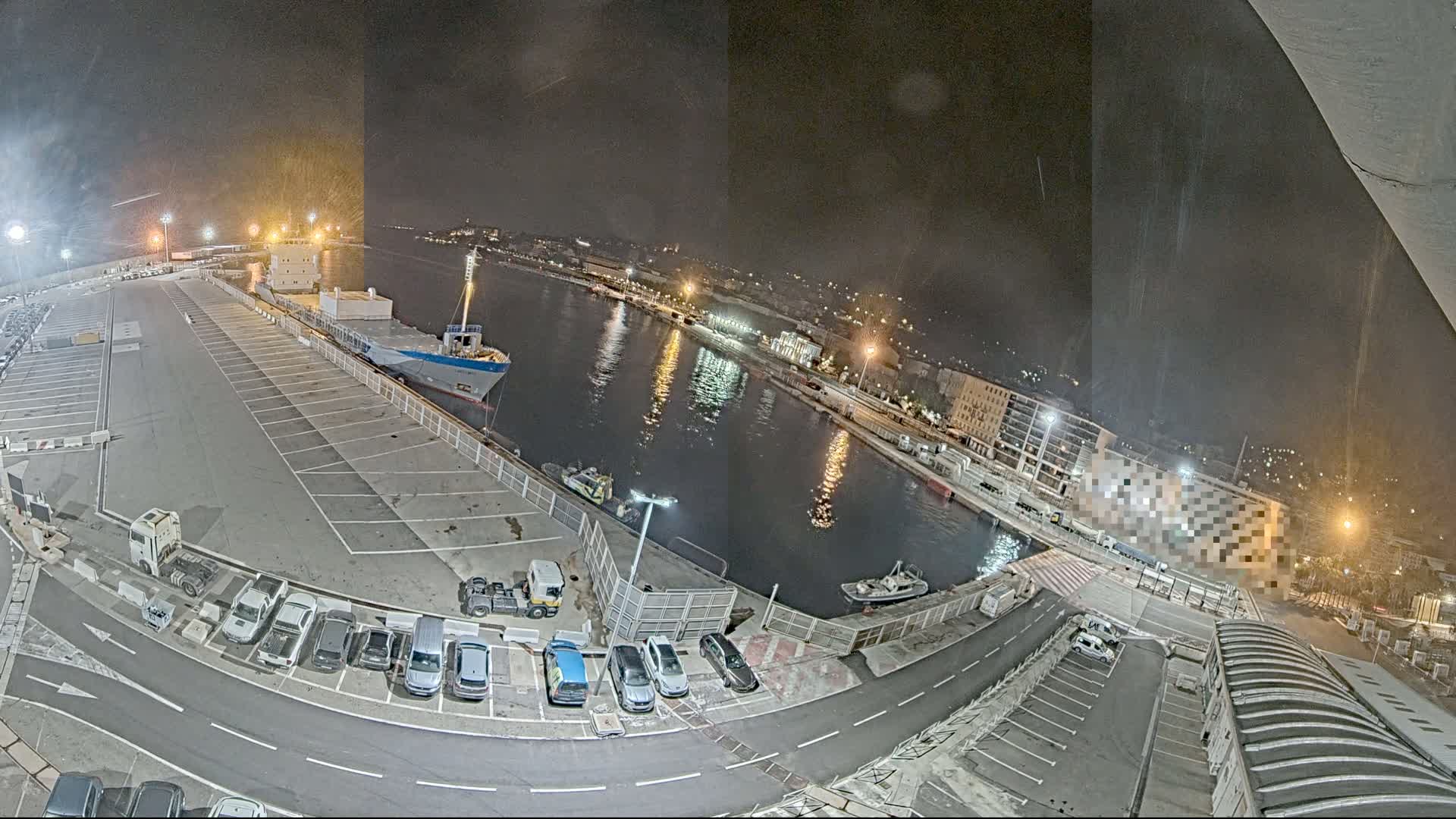 A nighttime view of a harbor shows a cargo ship docked, cars parked in a lot, and a calm body of water reflecting city lights.
