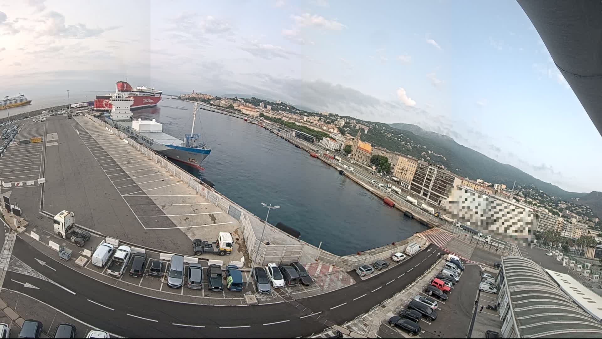 A mostly empty parking lot at a port with several ships docked, under a partly cloudy sky, overlooks a city nestled against mountains.