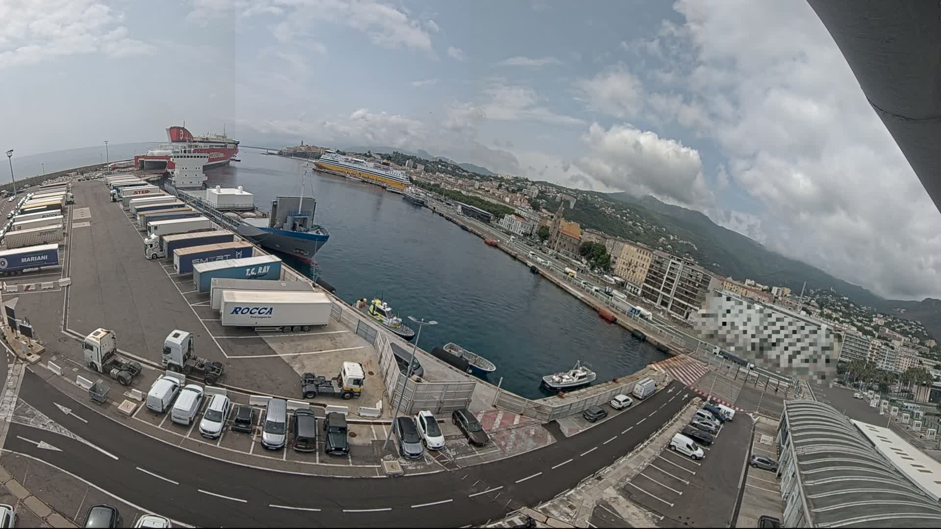 A port with several ferries and cargo ships, a parking lot filled with cars and trucks, and a city with mountains in the background under a partly cloudy sky.