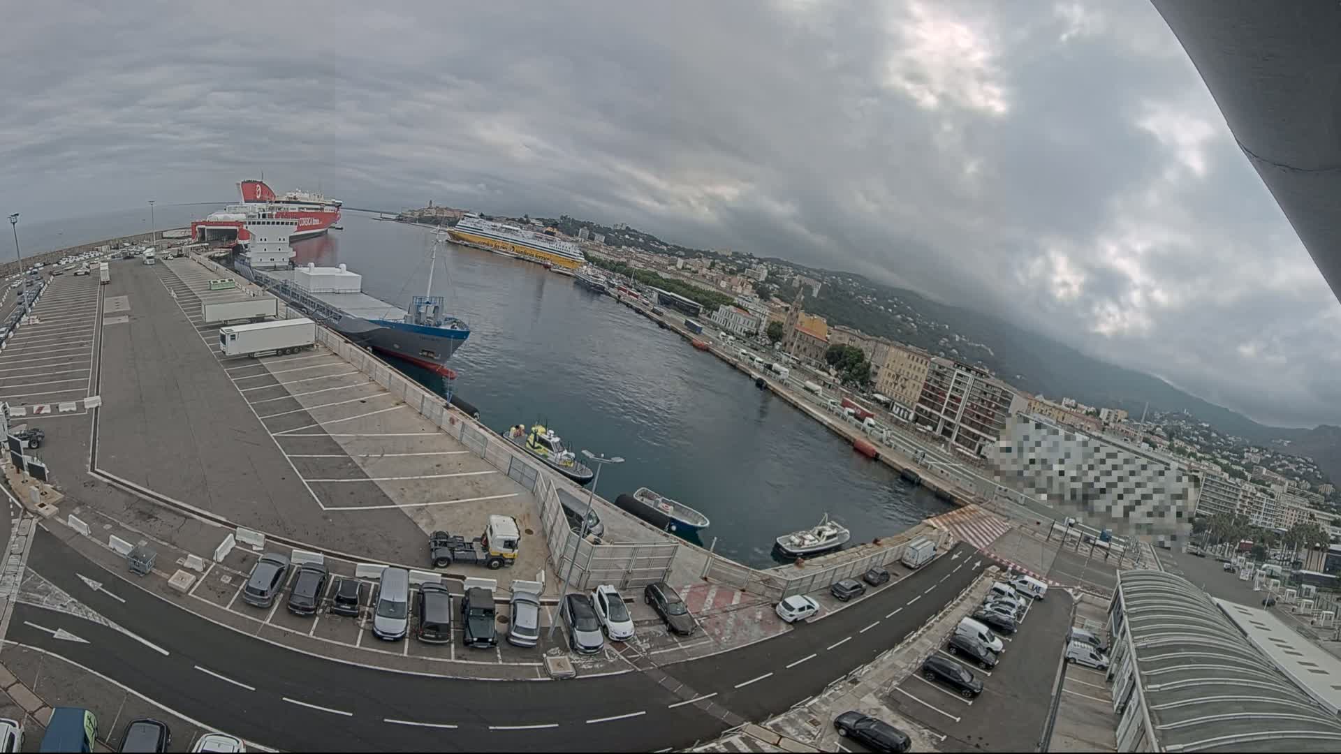 A harbor with several ships docked, cars parked in a lot, and buildings lining the waterfront under a cloudy sky.