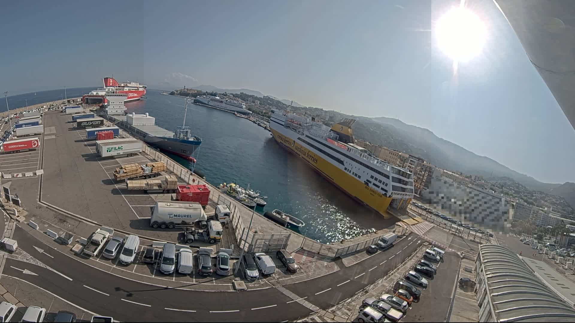 A sunny harbor scene shows several docked ferries, cargo ships, and numerous vehicles parked in a lot, with mountains visible in the distance.