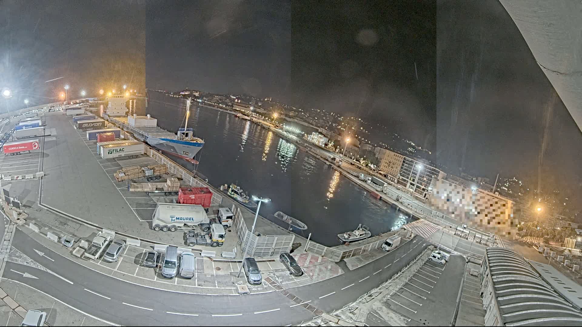 A nighttime aerial view of a harbor shows a cargo ship docked at a terminal with numerous trucks, containers, and cars in the surrounding parking lots, under a clear, dark sky.
