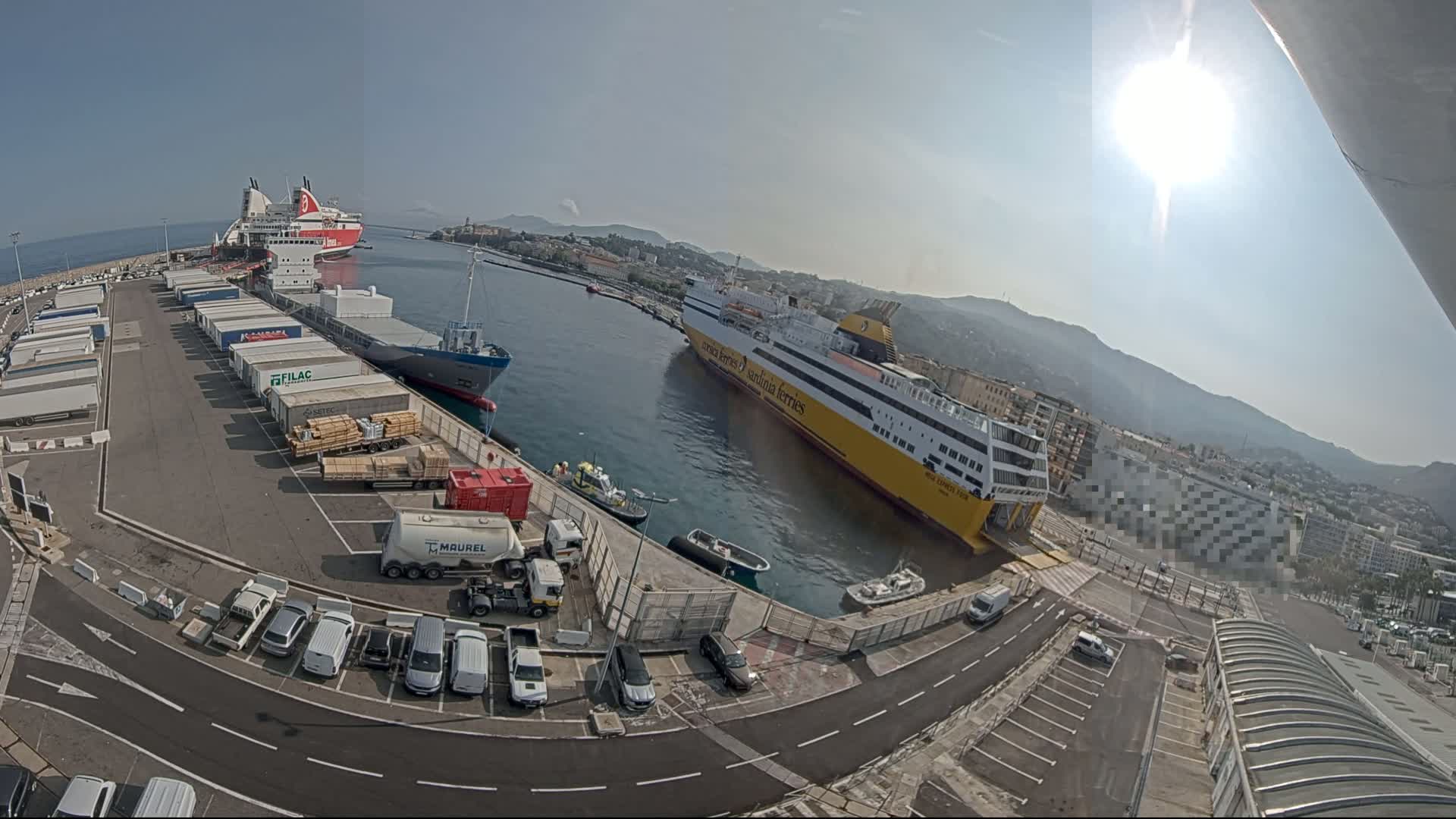 A sunny harbor scene shows two large ferries docked, with cargo containers and vehicles parked in the surrounding area.