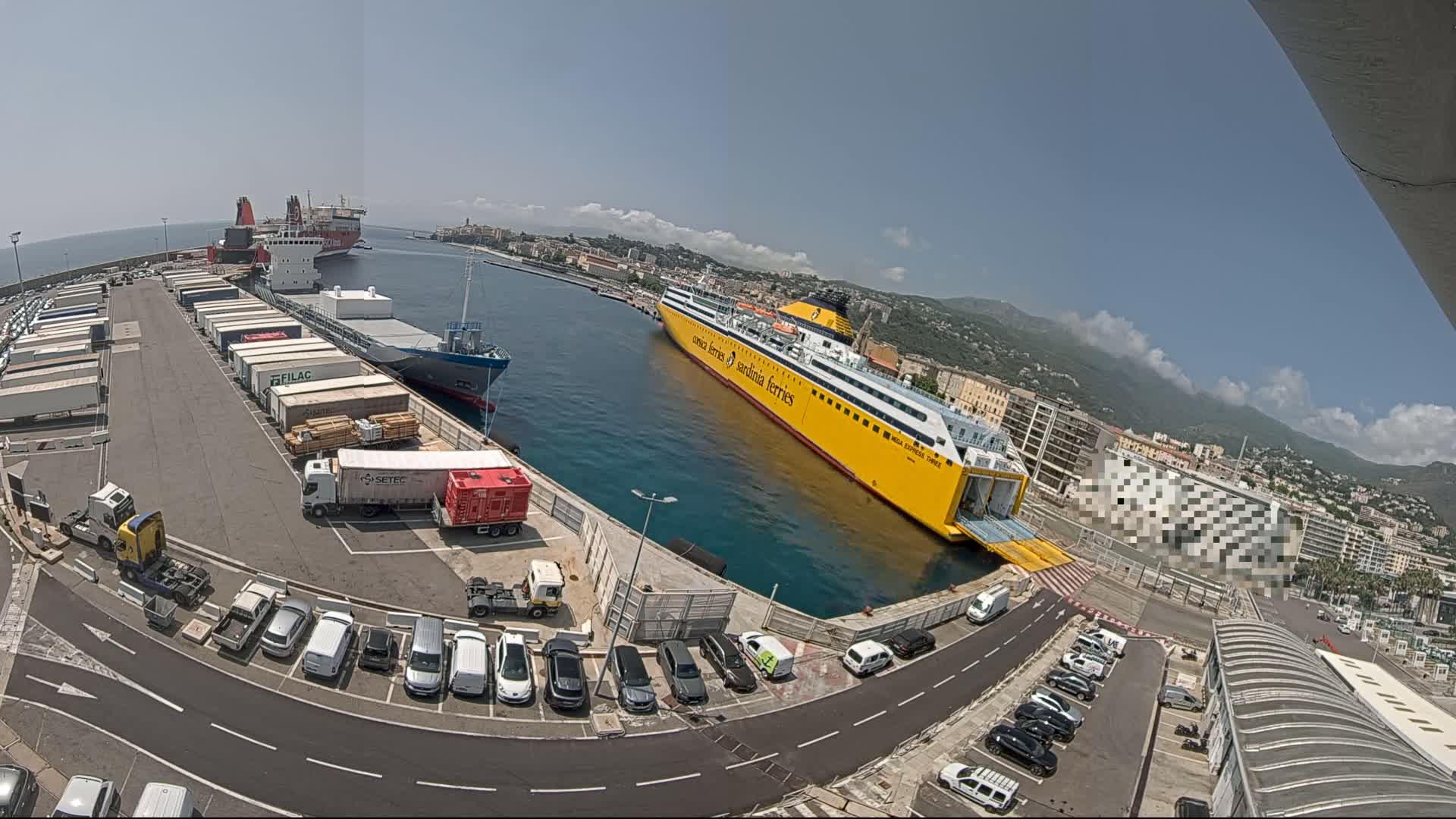A large yellow ferry is docked at a port on a sunny day, with several cargo ships, trucks, and cars visible in the surrounding area.