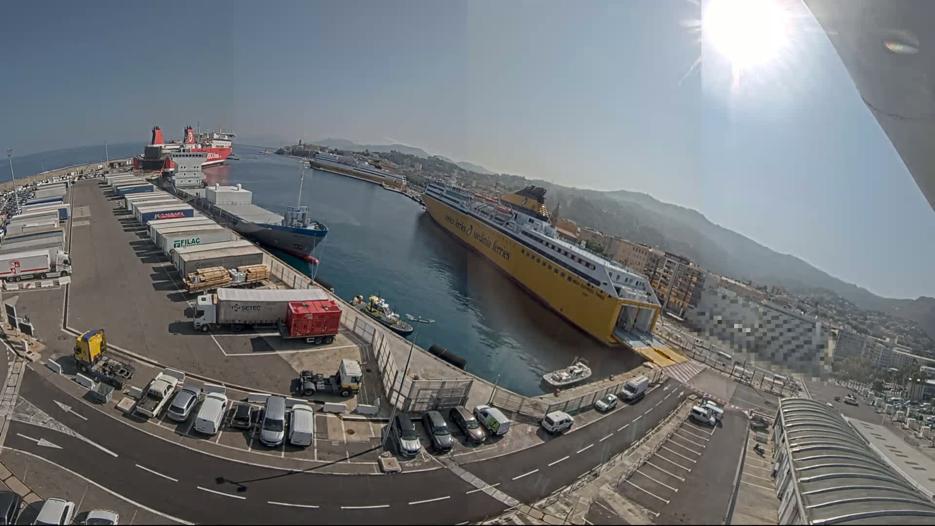 A sunny day at a harbor shows several ships docked, numerous cargo containers and trucks in a parking lot, and cars parked along roadways near buildings.