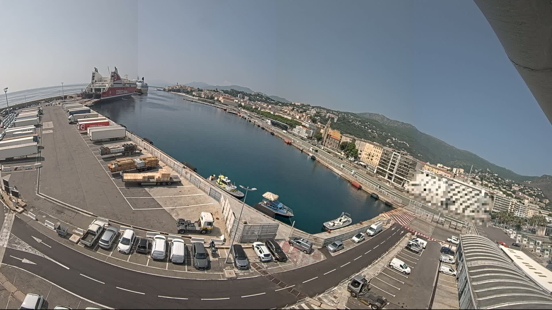A sunny day at a harbor shows a large ferry docked, cars parked in lots, smaller boats in the water, and a city in the background.