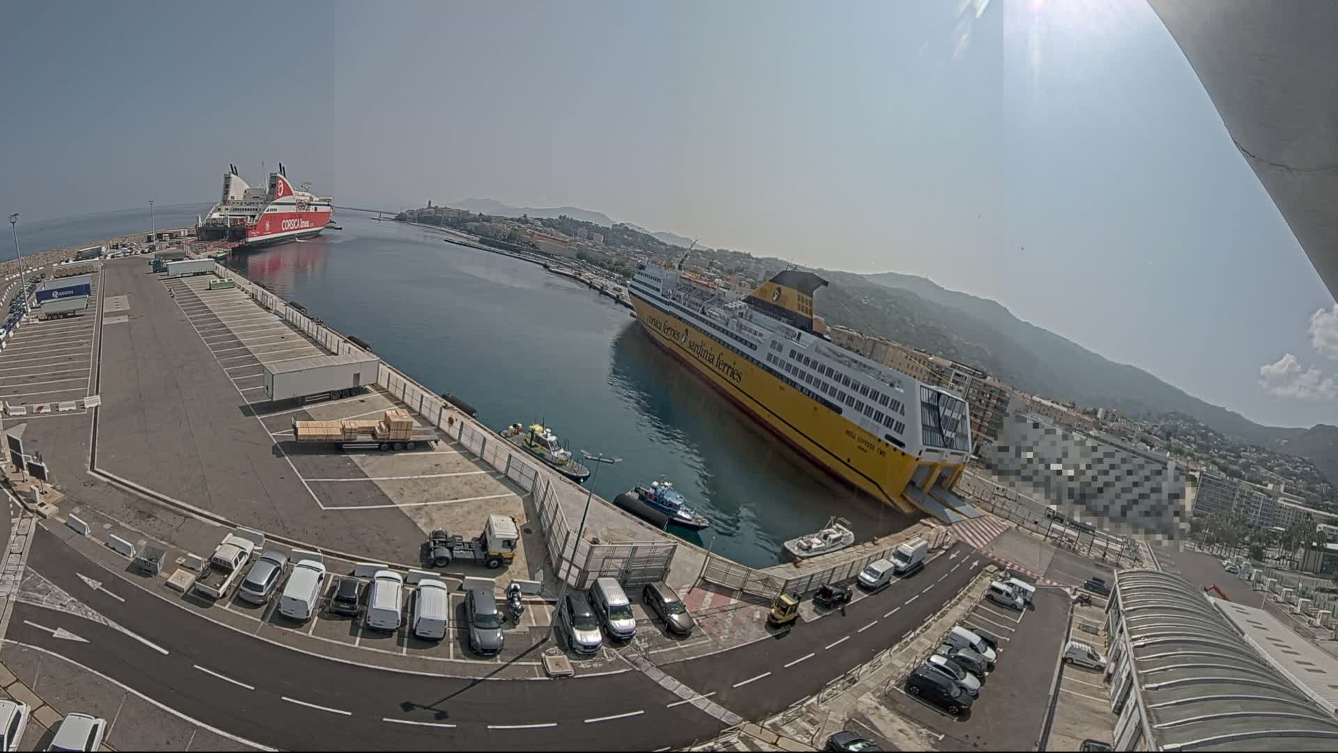 Two large ferries are docked at a port on a sunny day, with several cars parked in a lot nearby.