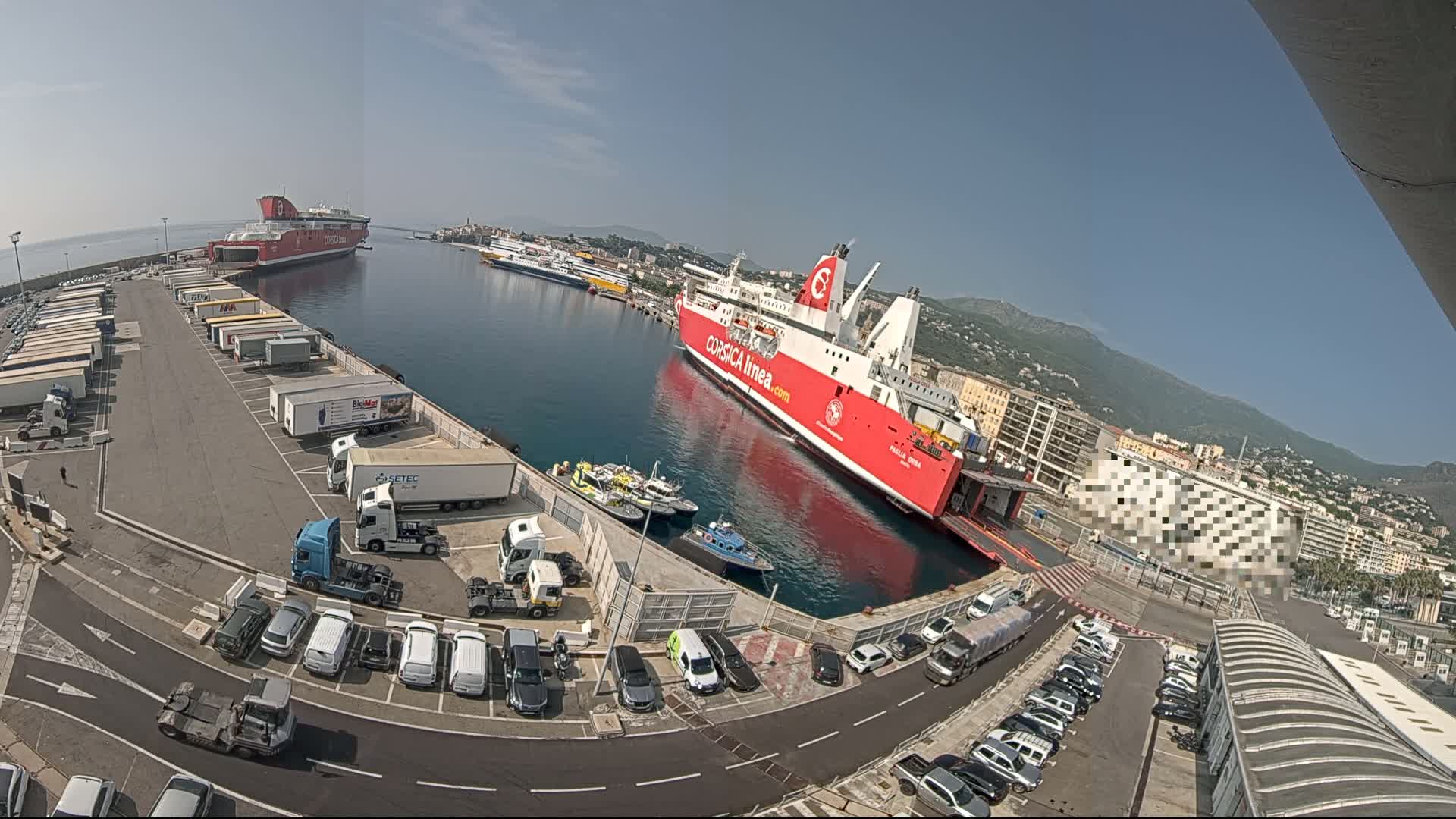 A large red ferry is docked at a port on a sunny day, with numerous cars and trucks parked in the surrounding areas.