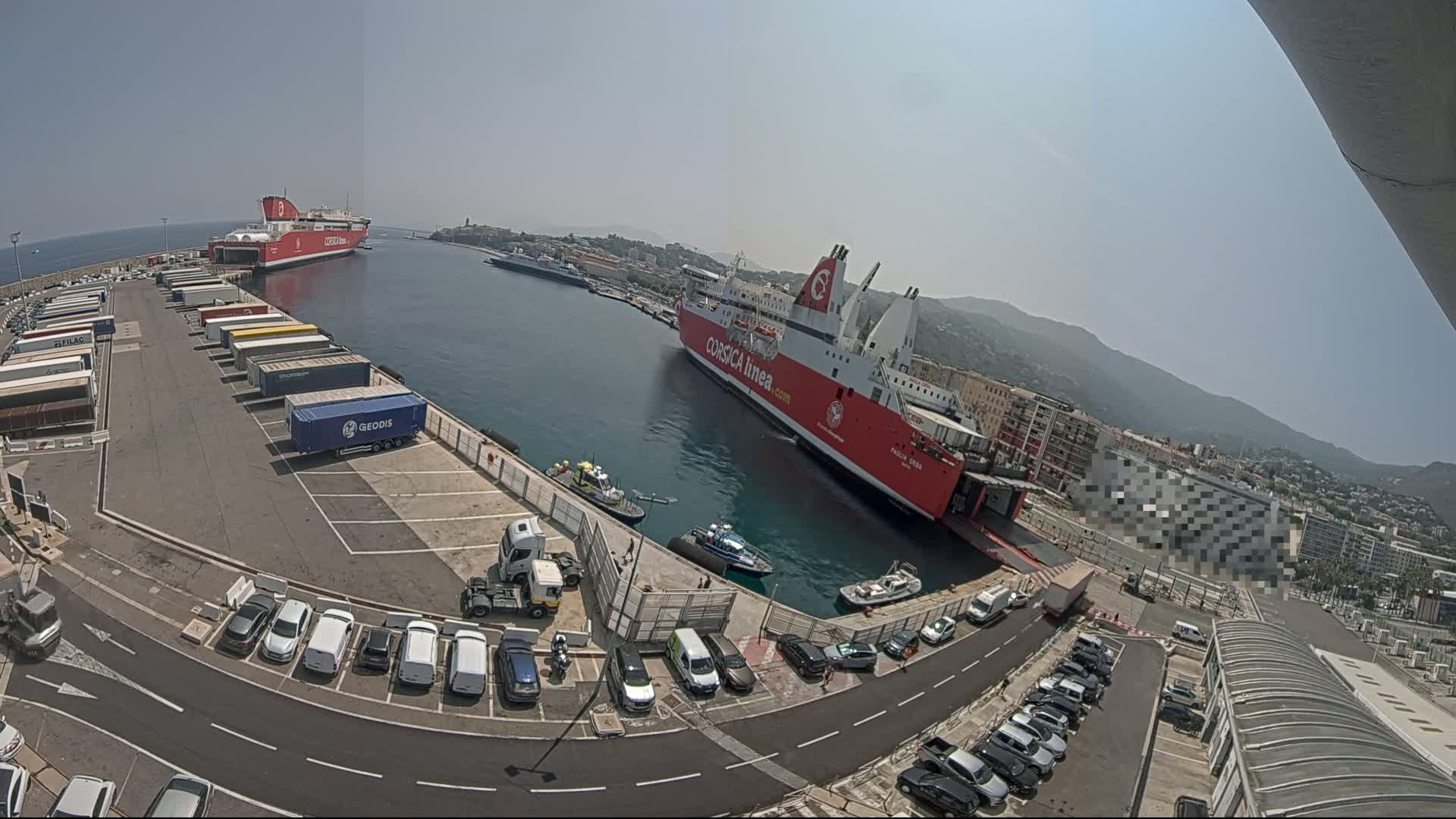 A sunny day at a port shows two large ferries docked, with numerous cars and trucks parked in the surrounding area, and mountains in the background.