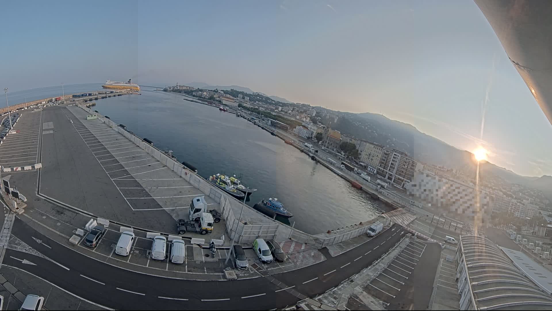 A high-angle, wide shot shows a port with a large empty parking lot, several small boats, a large ferry in the distance, and a city nestled against mountains, all under a bright, clear sunrise.