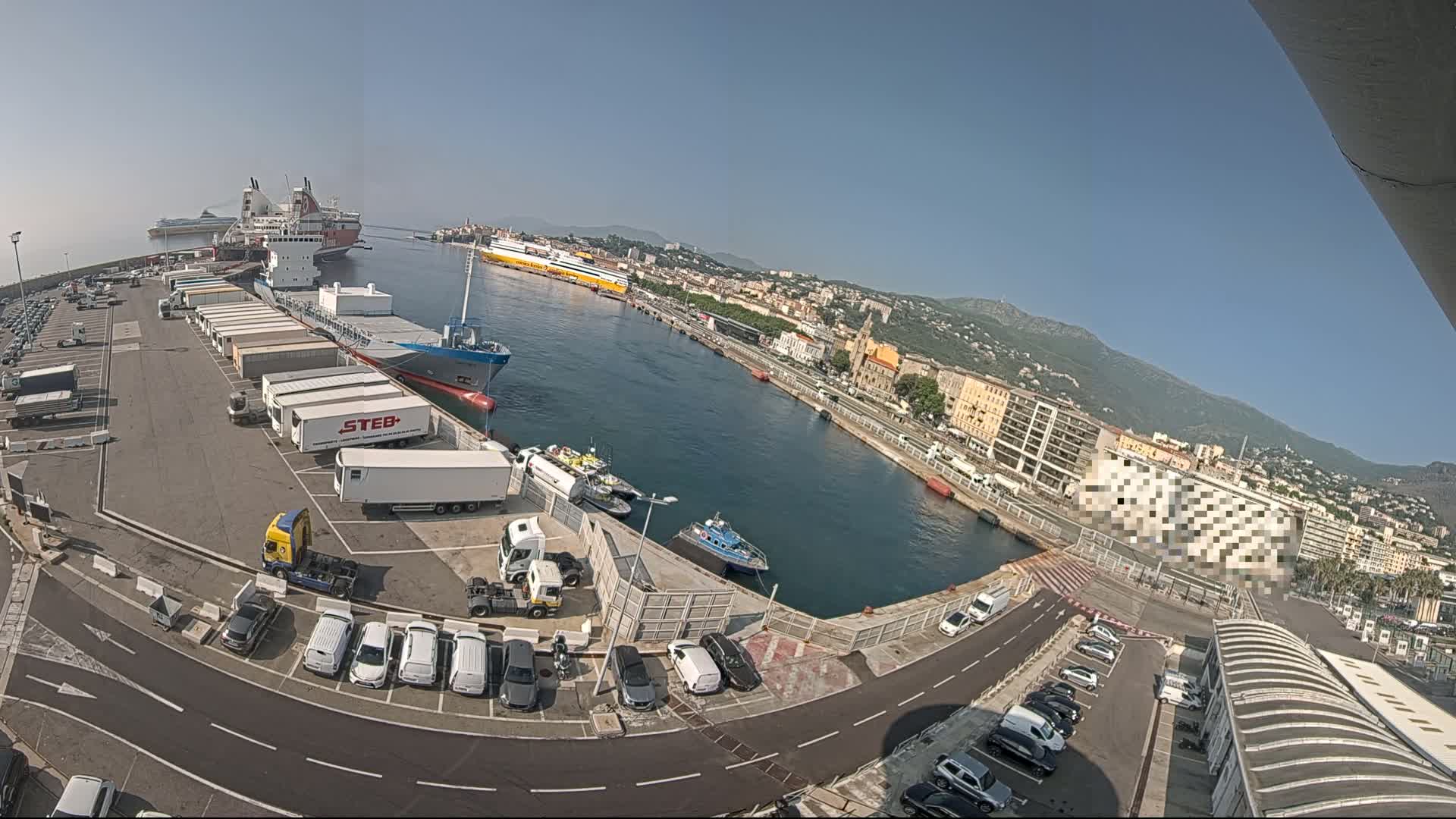 A harbor scene on a clear, sunny day shows several ships docked, numerous trucks and cars parked in lots, and a city in the background.