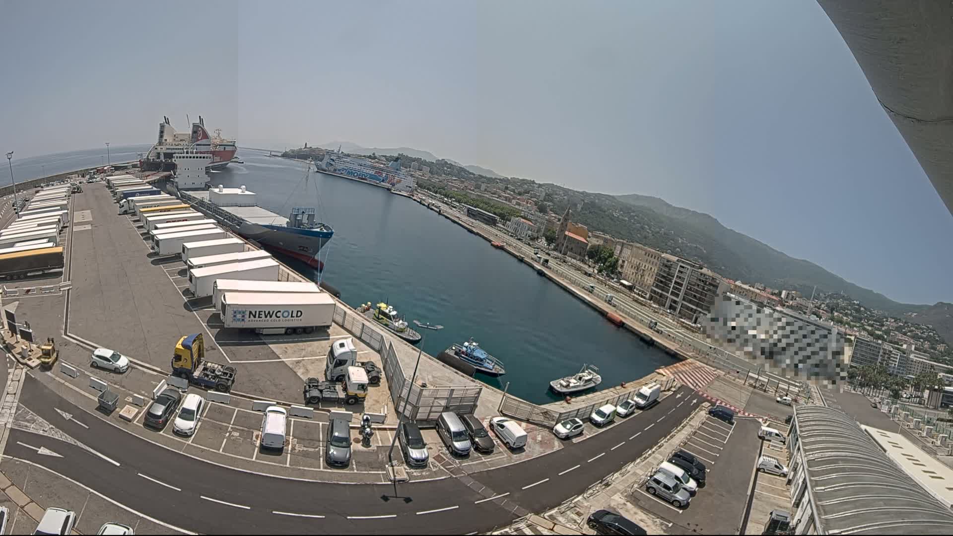 A harbor on a sunny day with several ships docked, numerous trailers parked on the dock, and cars in a parking lot alongside the water.