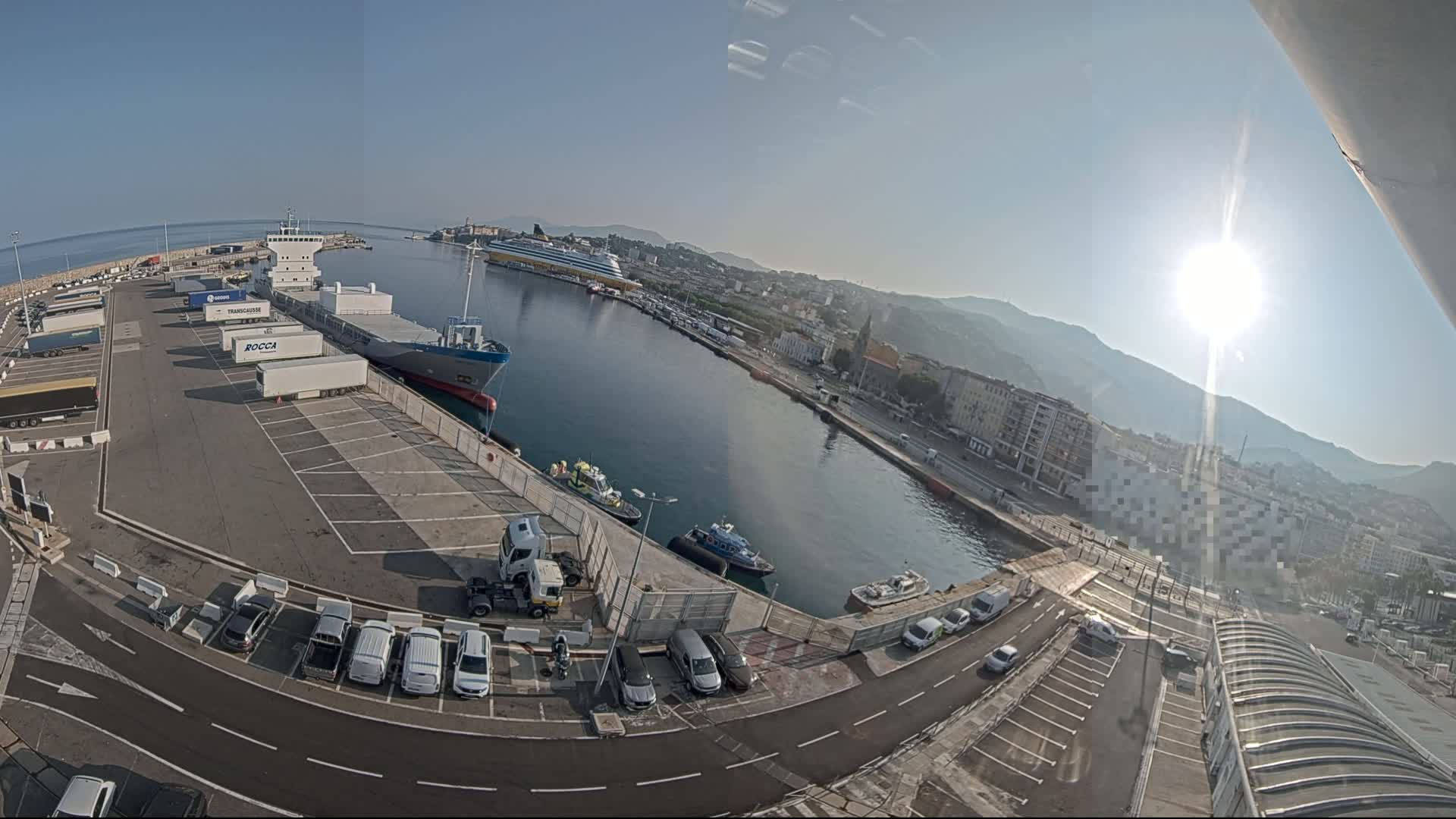 A sunny day at a harbor shows a cargo ship docked, numerous trailers and cars parked in lots, several smaller vessels in the water, and a city in the background with mountains beyond.
