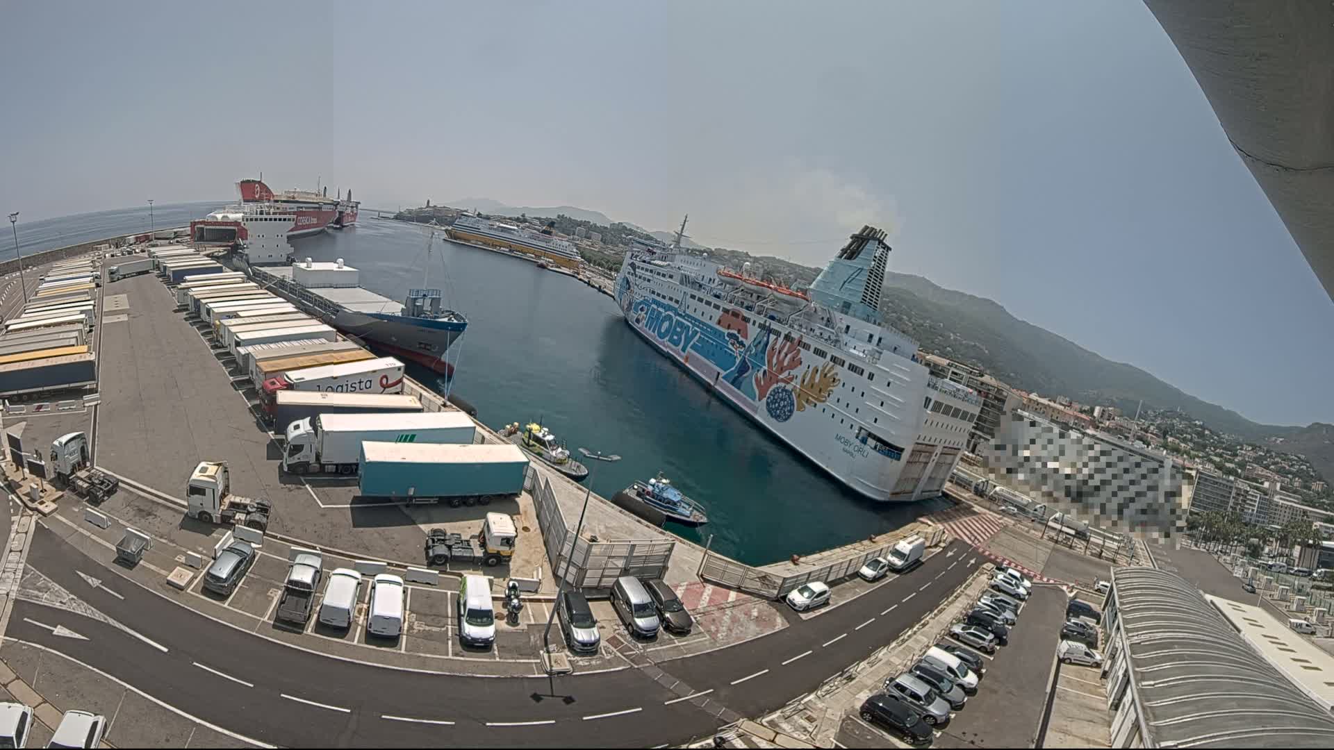 A harbor on a sunny day with several large ships docked, numerous trucks and cars parked in a lot, and smaller vessels in the water.