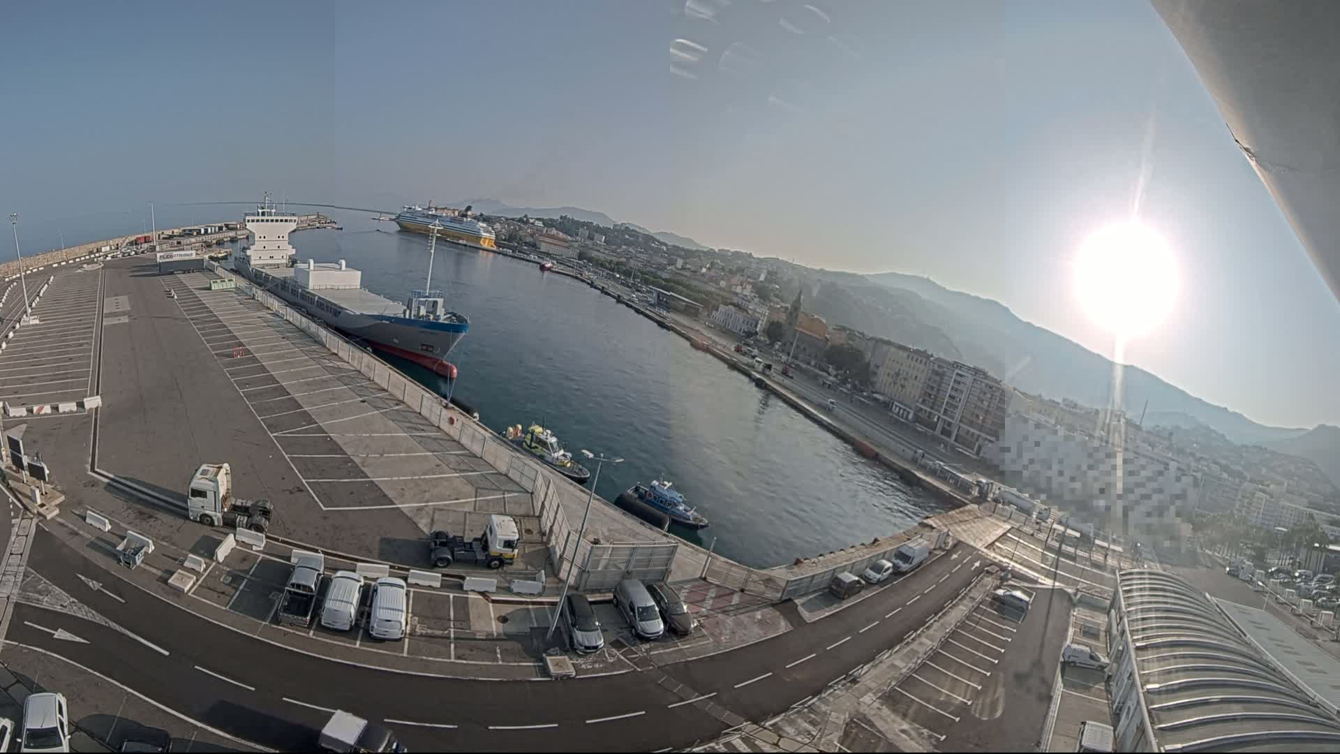 A sunny harbor scene shows a large cargo ship docked near several smaller boats, a large parking lot, and a city on a hillside in the background.