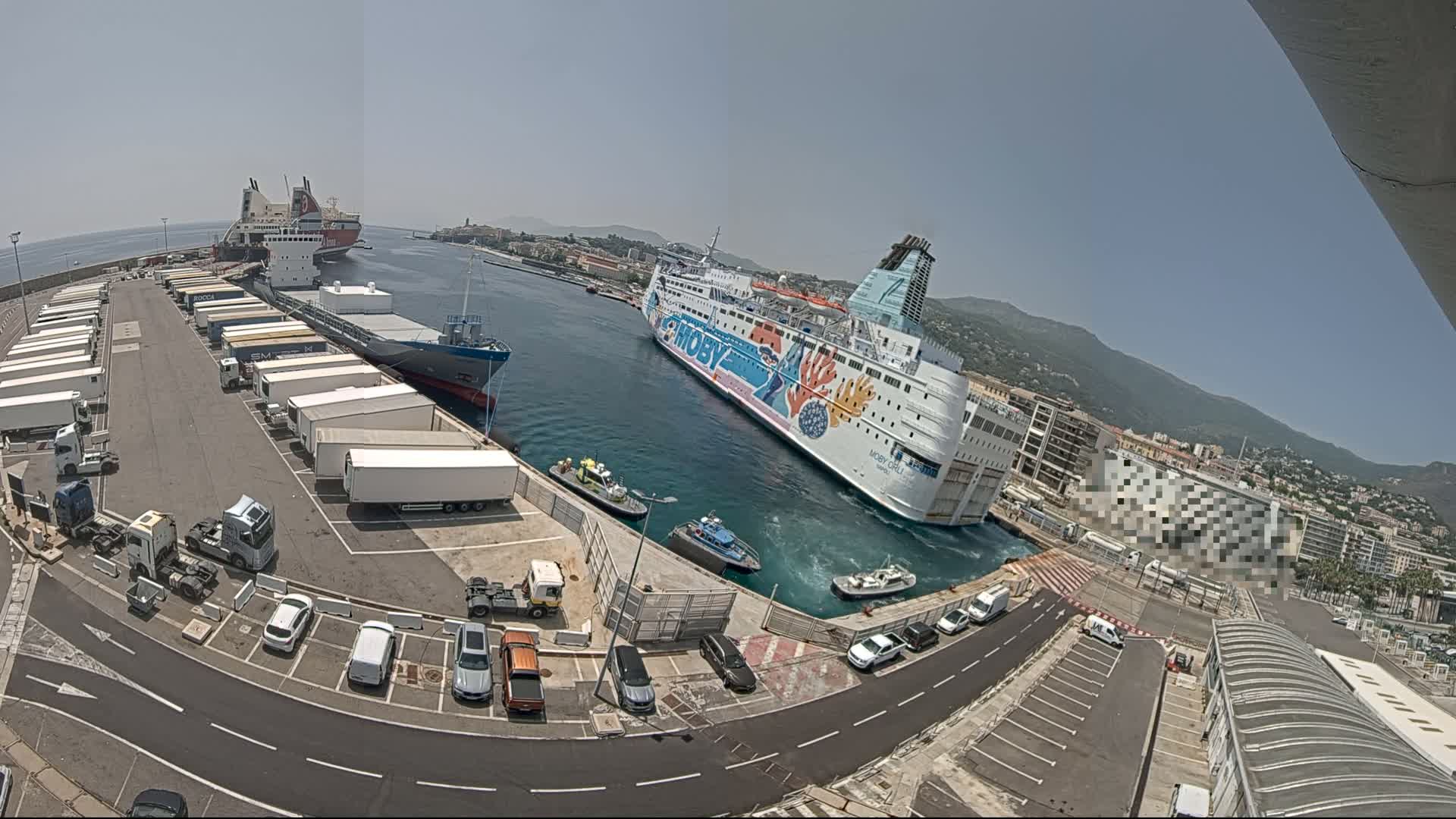 A large ferry is docked at a port on a sunny day, with numerous trucks and cars parked nearby, along with smaller support vessels.