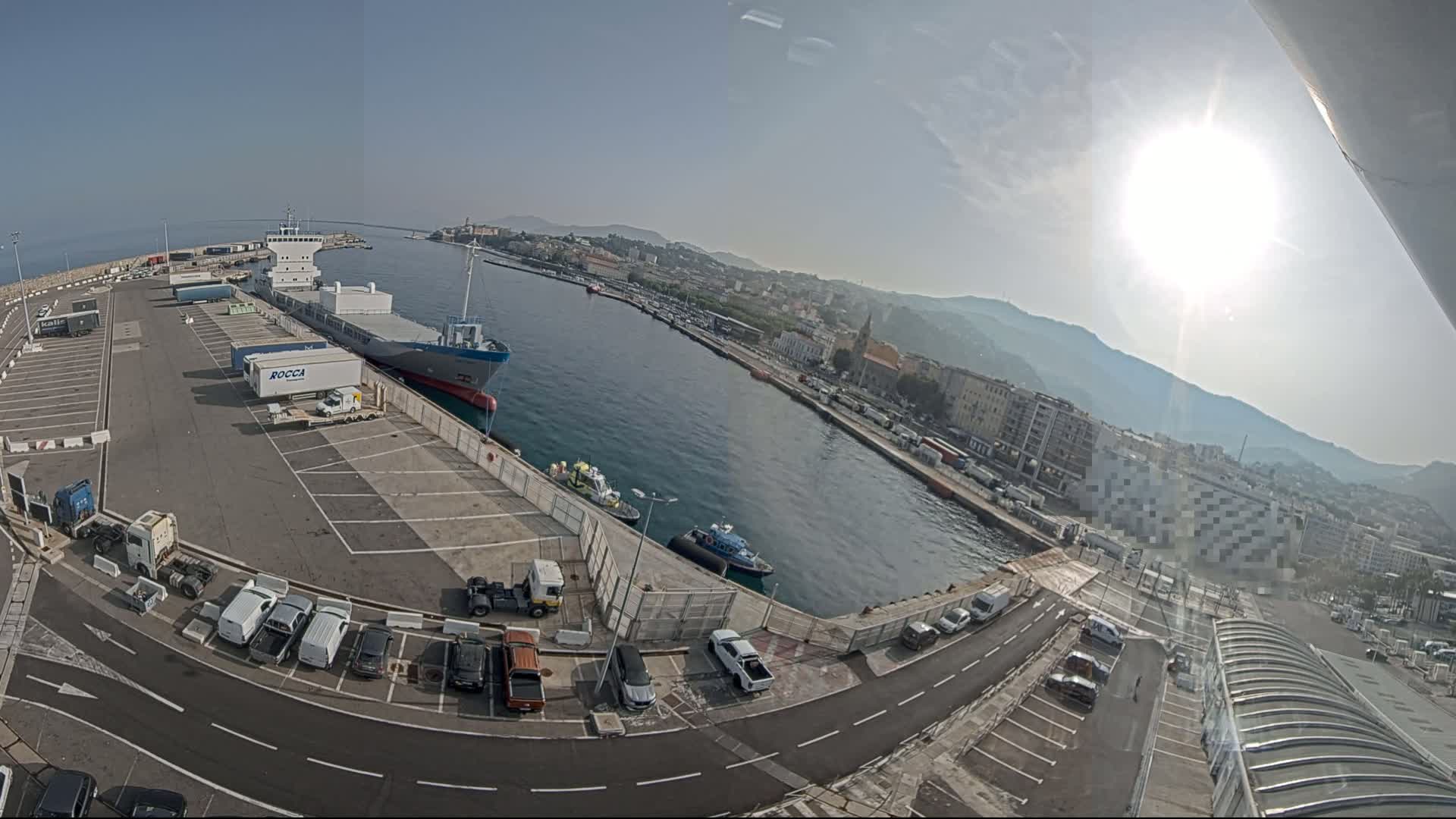 A sunny day at a harbor shows a cargo ship docked, numerous vehicles parked in a lot, and a city in the background across a body of water.
