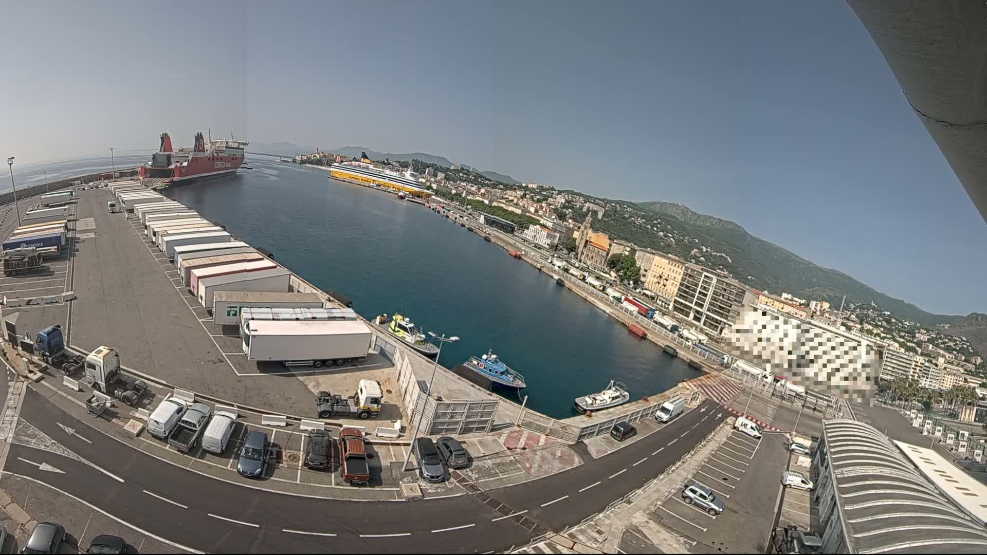 A high-angle, wide shot on a sunny day shows a harbor with two ferries, numerous trucks and cars in a parking lot, and a city in the background.