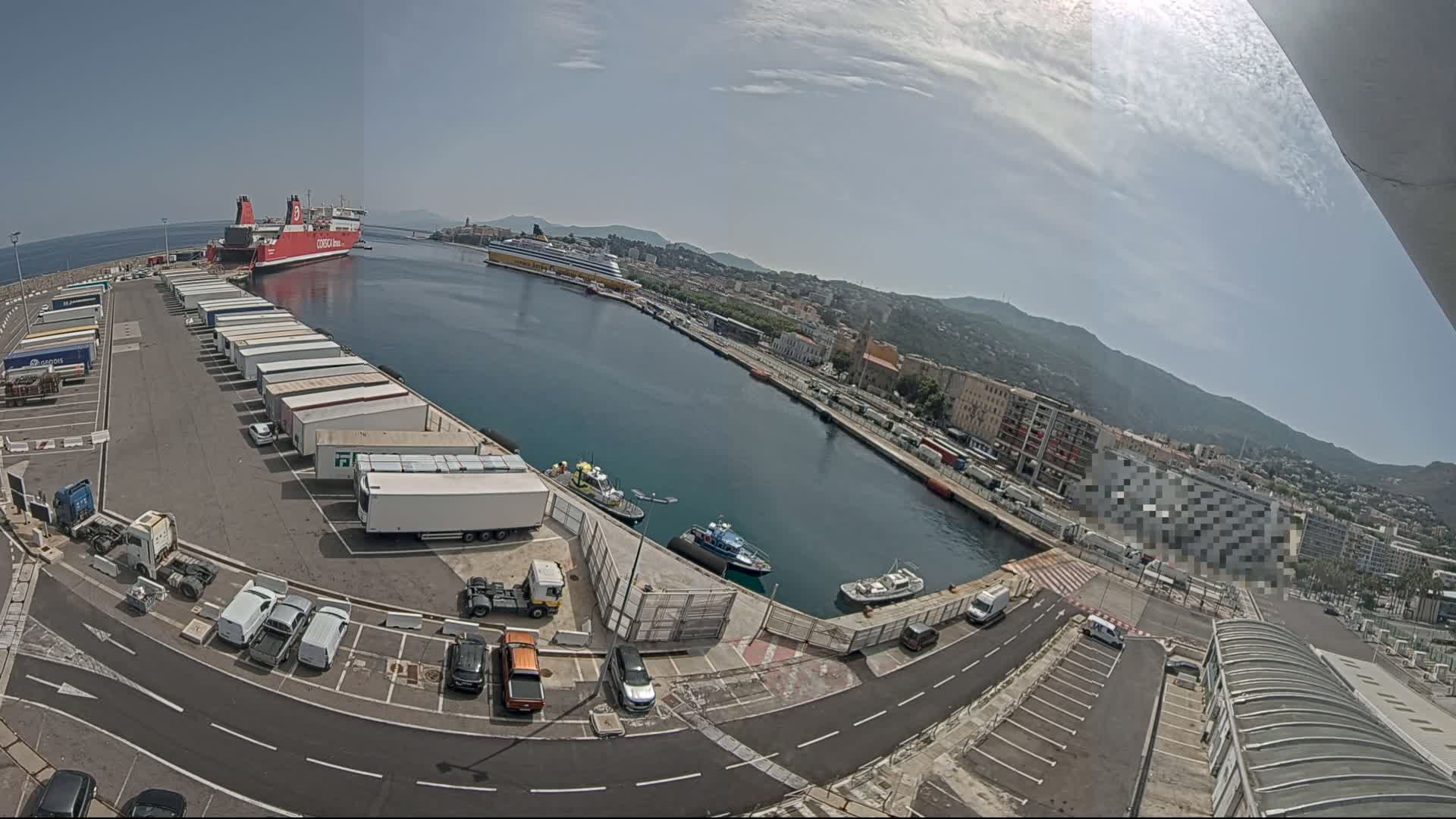 A harbor on a sunny day with a large red ferry, several smaller boats, numerous trucks parked in a lot, and a city in the background.
