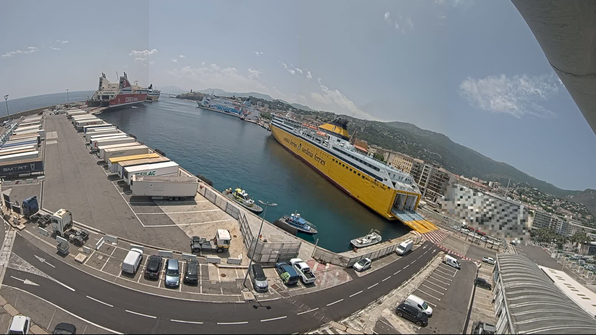A sunny day at a busy port shows several ferries docked, numerous cargo trucks and cars parked, and smaller boats in the water.