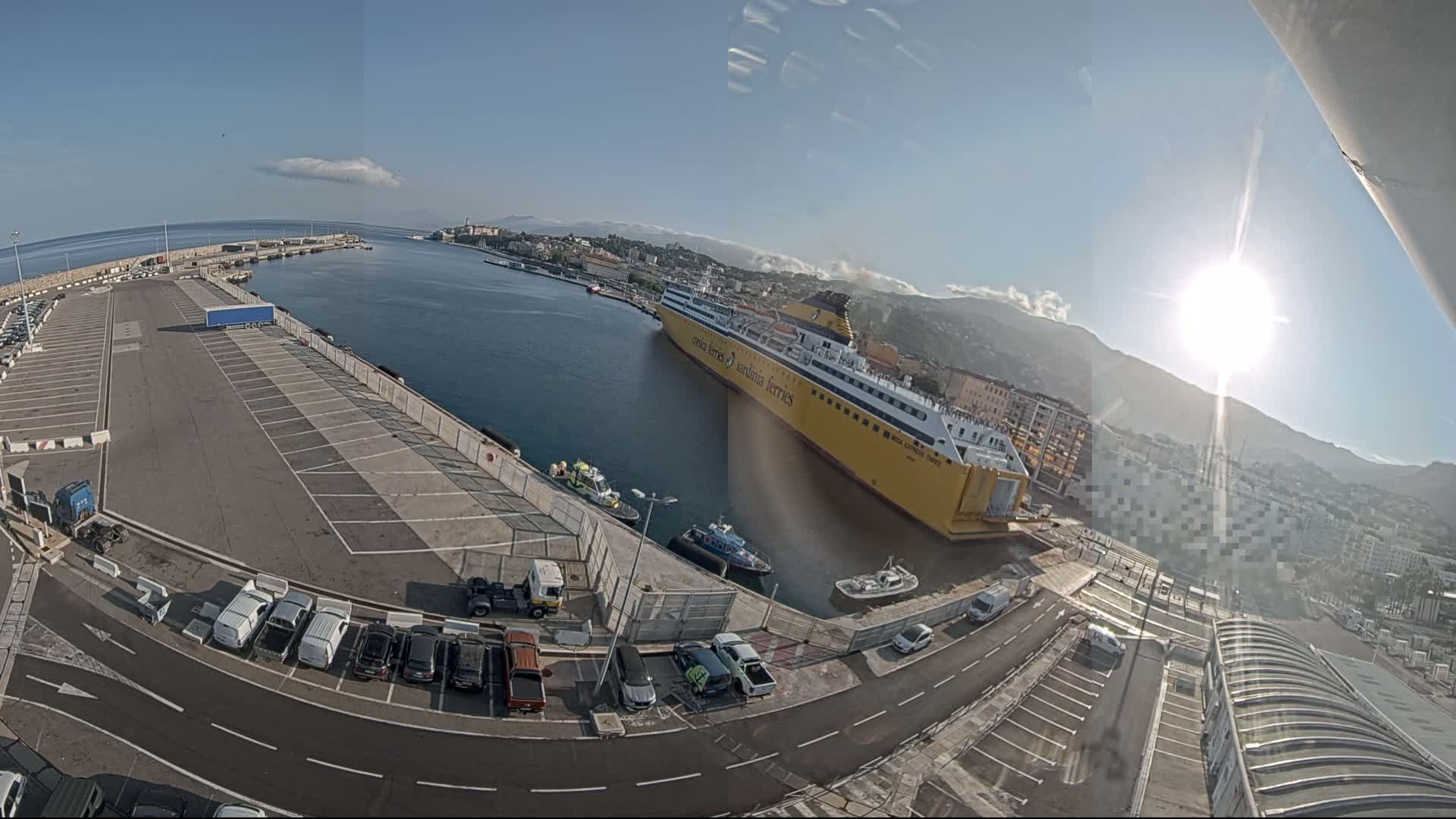 A large yellow ferry is docked at a port on a sunny day with clear skies and mountains in the background.