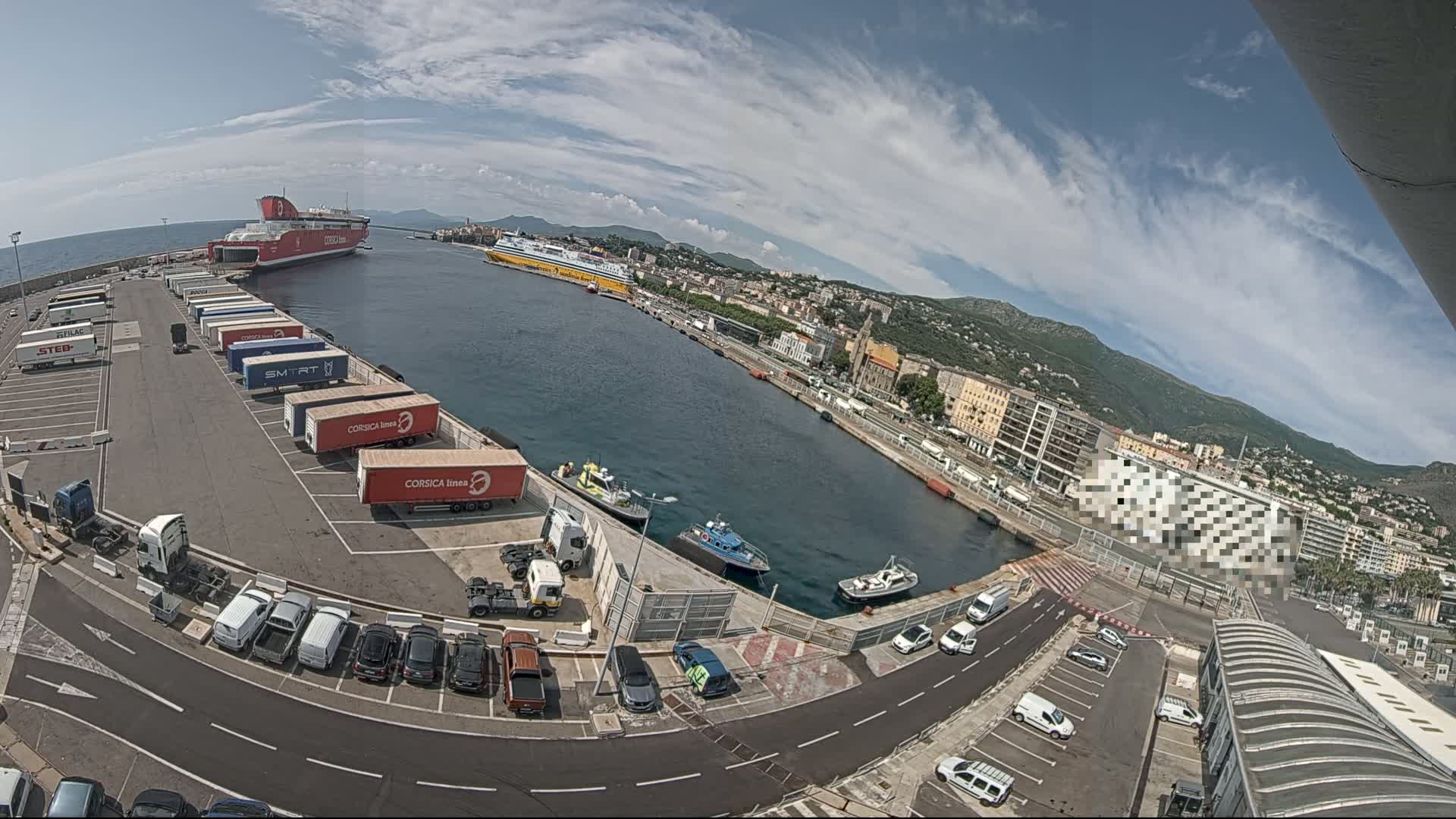 A ferry port on a sunny day with a partly cloudy sky, showing two large ferries docked, numerous trucks and cars parked, and several smaller boats in the water.