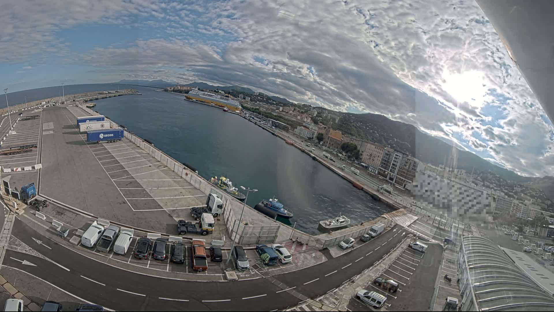 A partly sunny day at a harbor shows a large ferry, several smaller boats, numerous vehicles parked in a lot, and a city in the background.