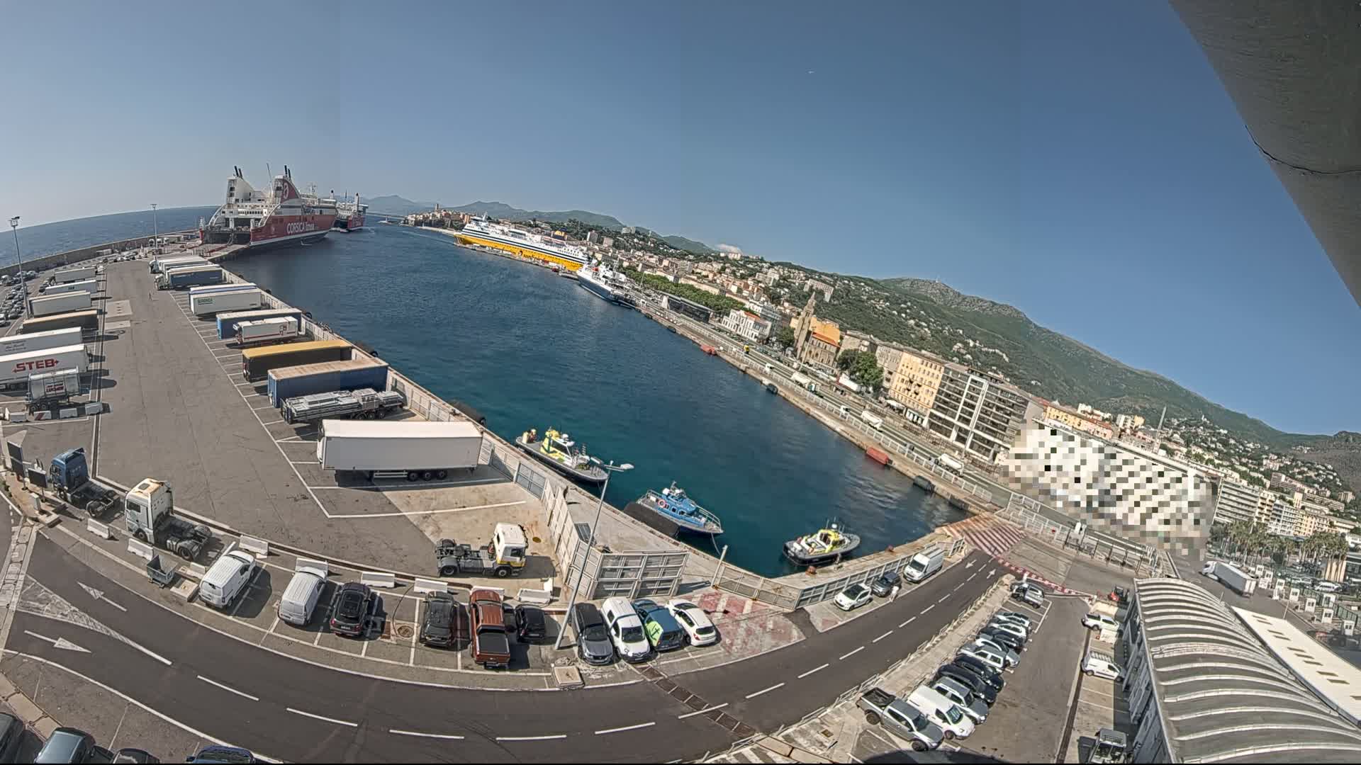 A sunny day at a harbor shows several docked ferries, numerous trucks and cars parked in lots, and several small boats in the water.