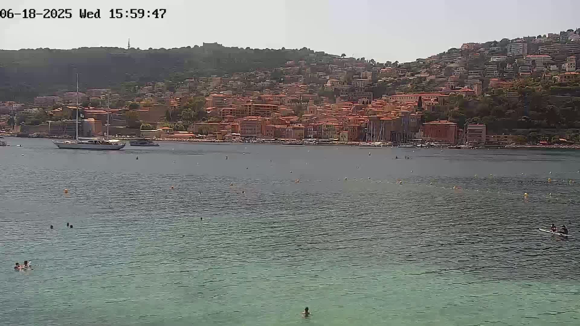A large sailboat is anchored in calm water in front of a hillside town on a sunny day.